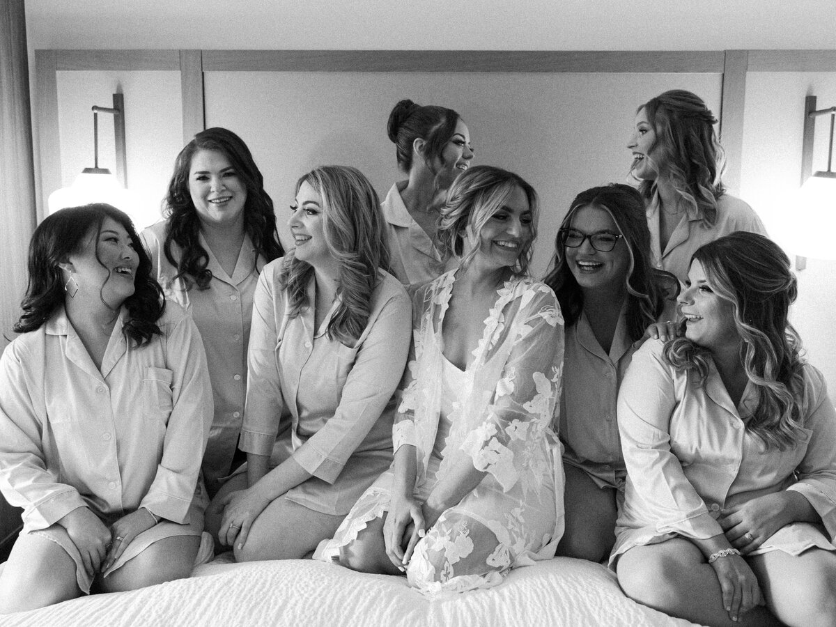 A bride laughs with her bridesmaids on a hotel room bed as she gets ready for her wedding