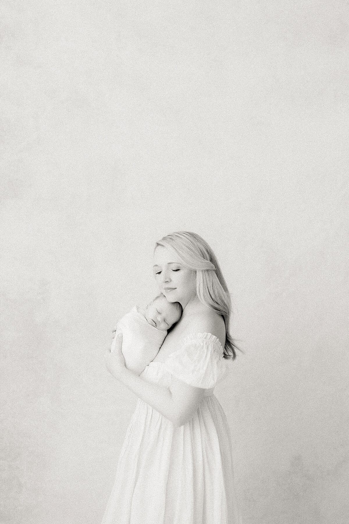 Black and white newborn portrait of a mother standing in a DFW photography studio while she holds her newborn baby girl to her chest.
