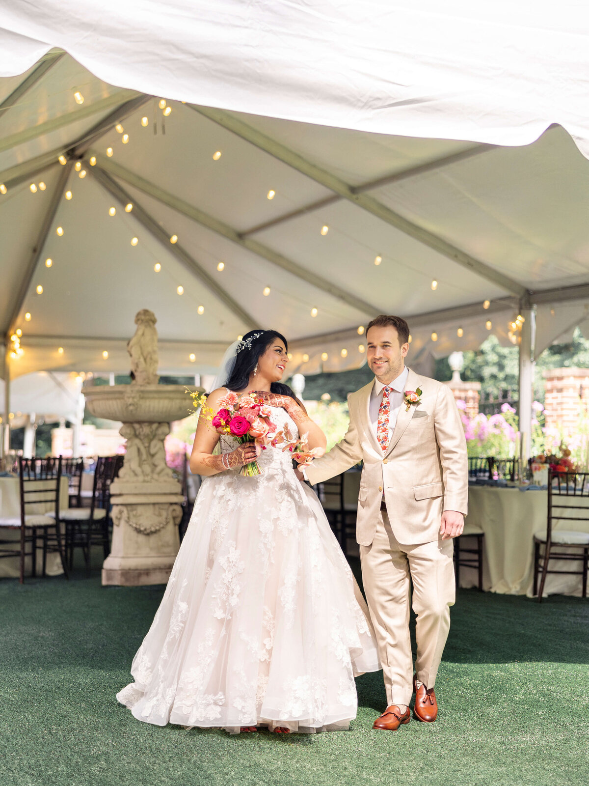 A bride and groom smile and walk under a white tent decorated with string lights. The bride holds a bouquet of pink and orange flowers, wearing a white gown, while the groom is in a beige suit with a floral tie. Tables and chairs are in the background.