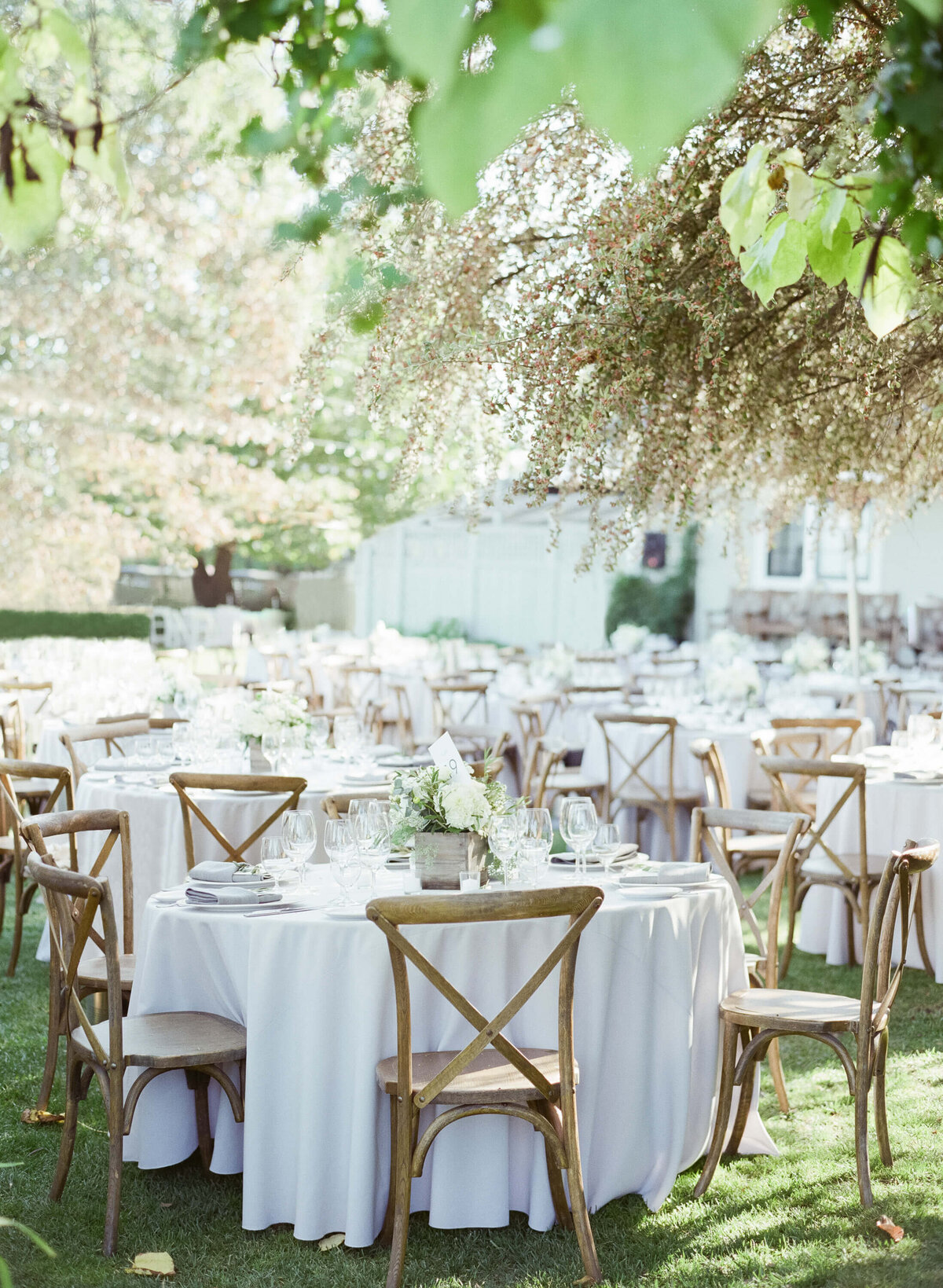 Exquisite arrangement of outdoor wedding reception tables with vintage wooden chairs and white tablecloths beside a beautiful scenery of green garden floor and white flowers.