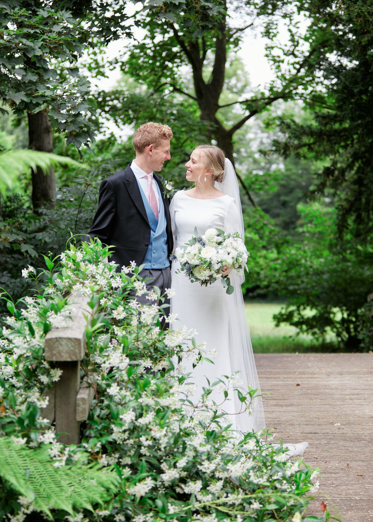 bride in beautiful satin wedding dress and groom in suit with pink tie and blue vest posing on the wooden bridge among greenery