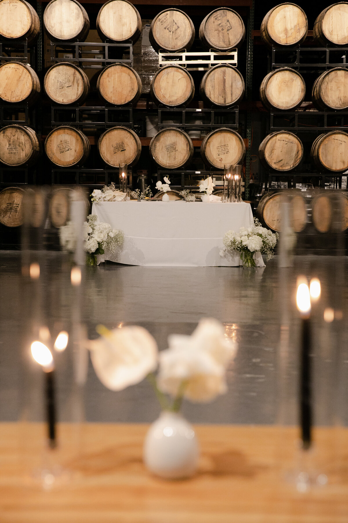 Minimalist wedding centerpieces are displayed with black candles and a white vase with white lilies out of focus and beer barrels at Moody Tongue Brewery in the background.