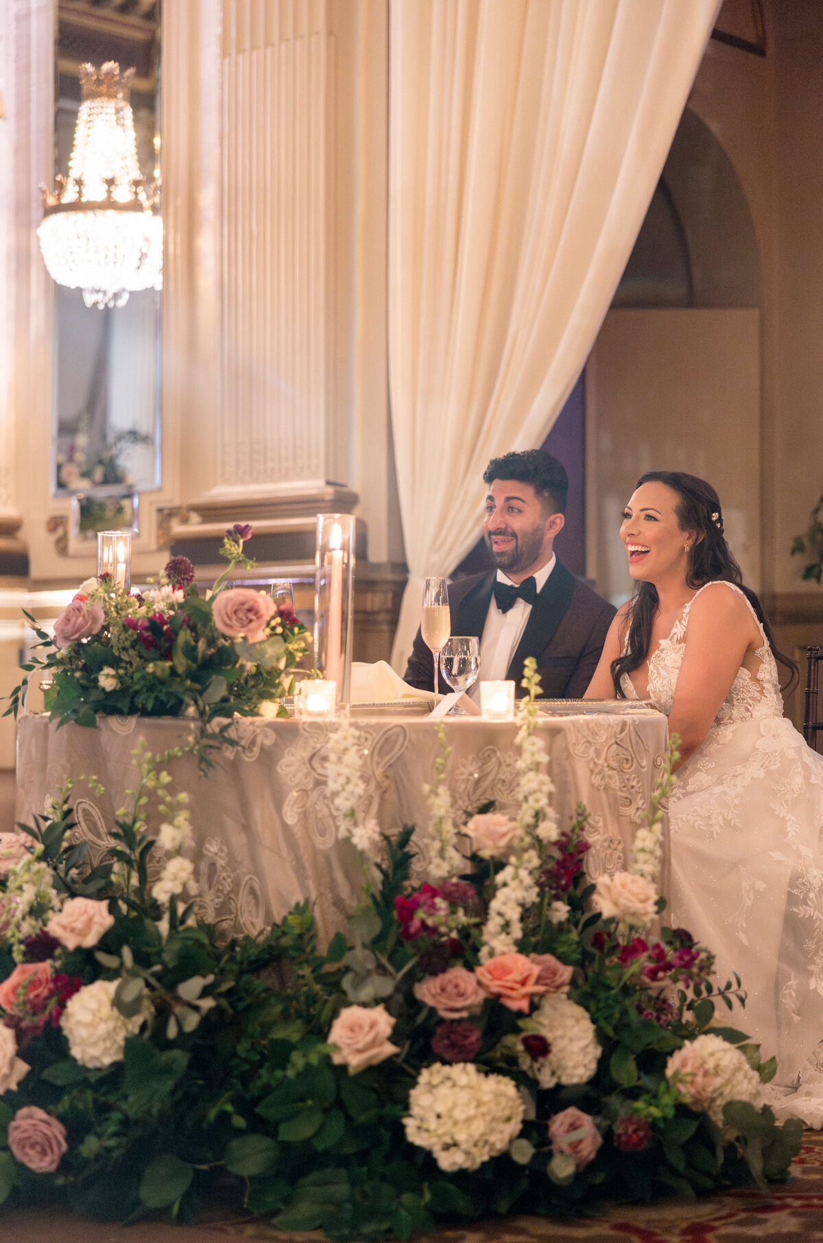 A bride and groom sit at a decorated table, smiling and laughing. The table is adorned with elegant flowers in shades of pink and white, and tall candles. A chandelier hangs above in a warmly lit venue.