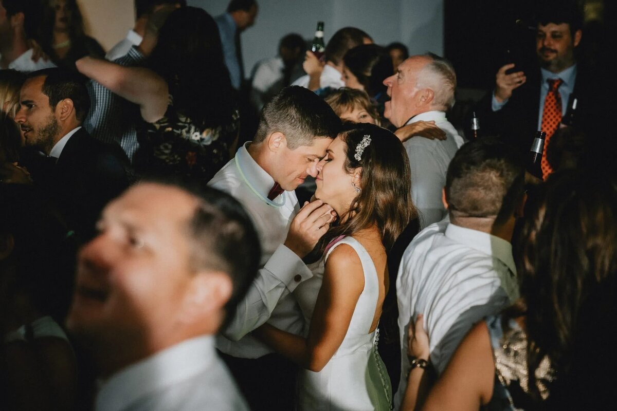 In the midst of a bustling wedding reception, a bride and groom share a tender, private moment, their faces close together, surrounded by guests.