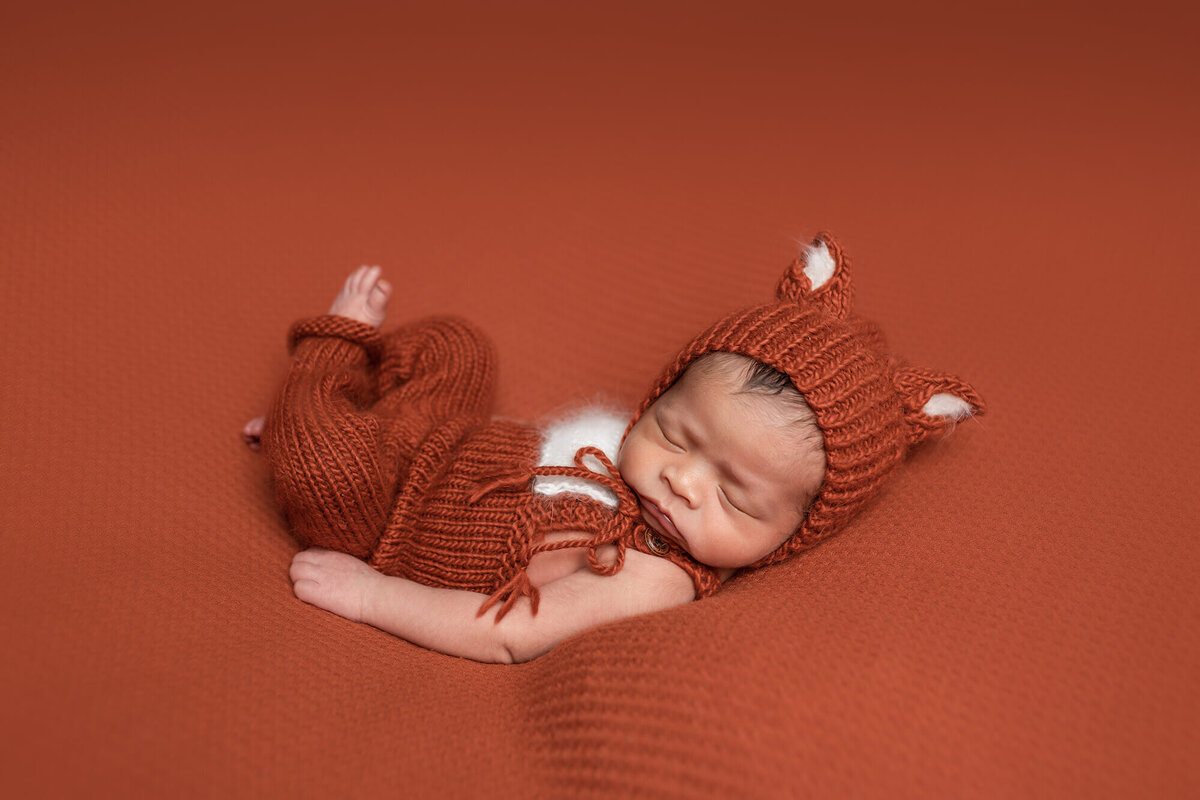 A newborn baby in a knitted, orange fox outfit sleeps on an orange blanket