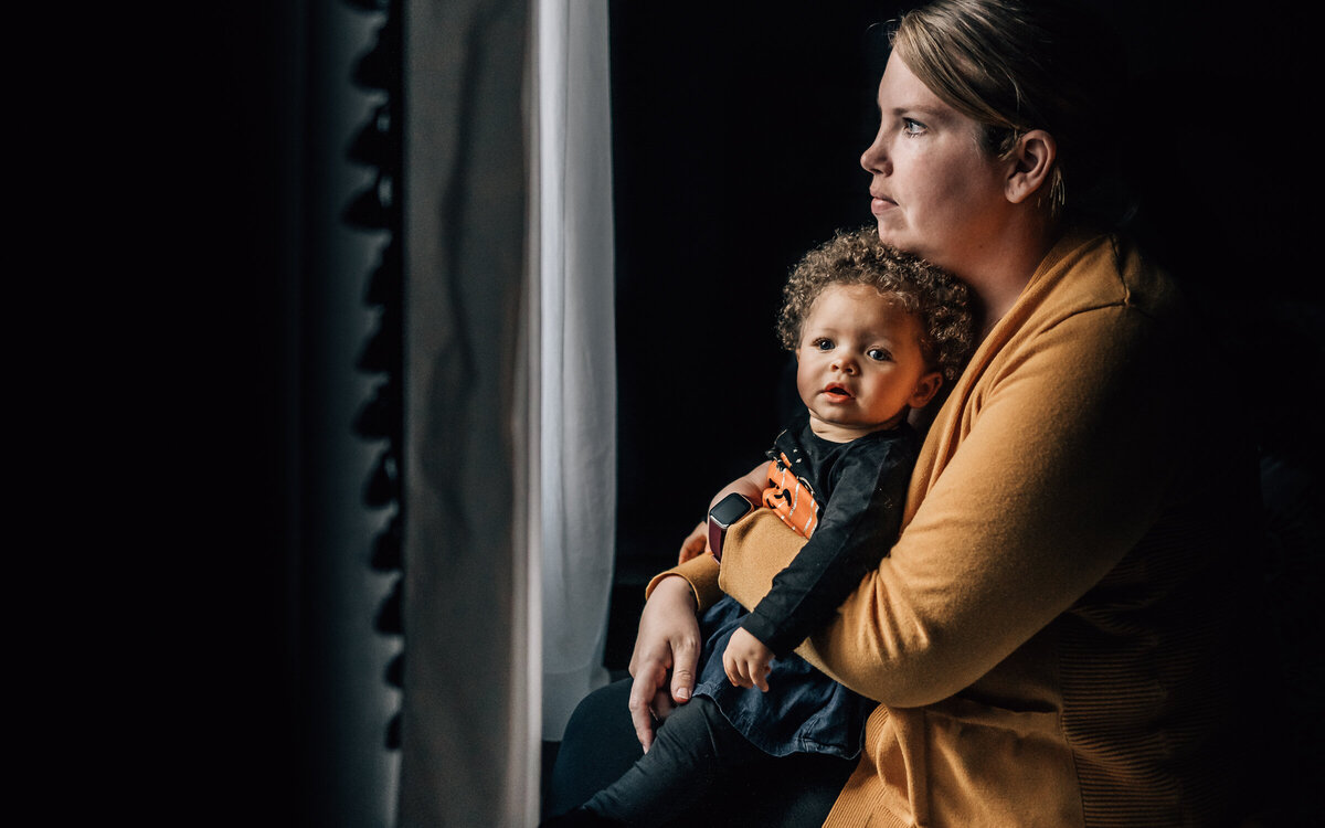 A mom, illuminated dramatically by window light, gazes outside deep in though while her child locks eyes with the camera.