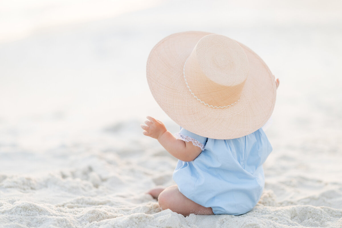 A baby sitting in the sand with a big hat on in Alys Beach.