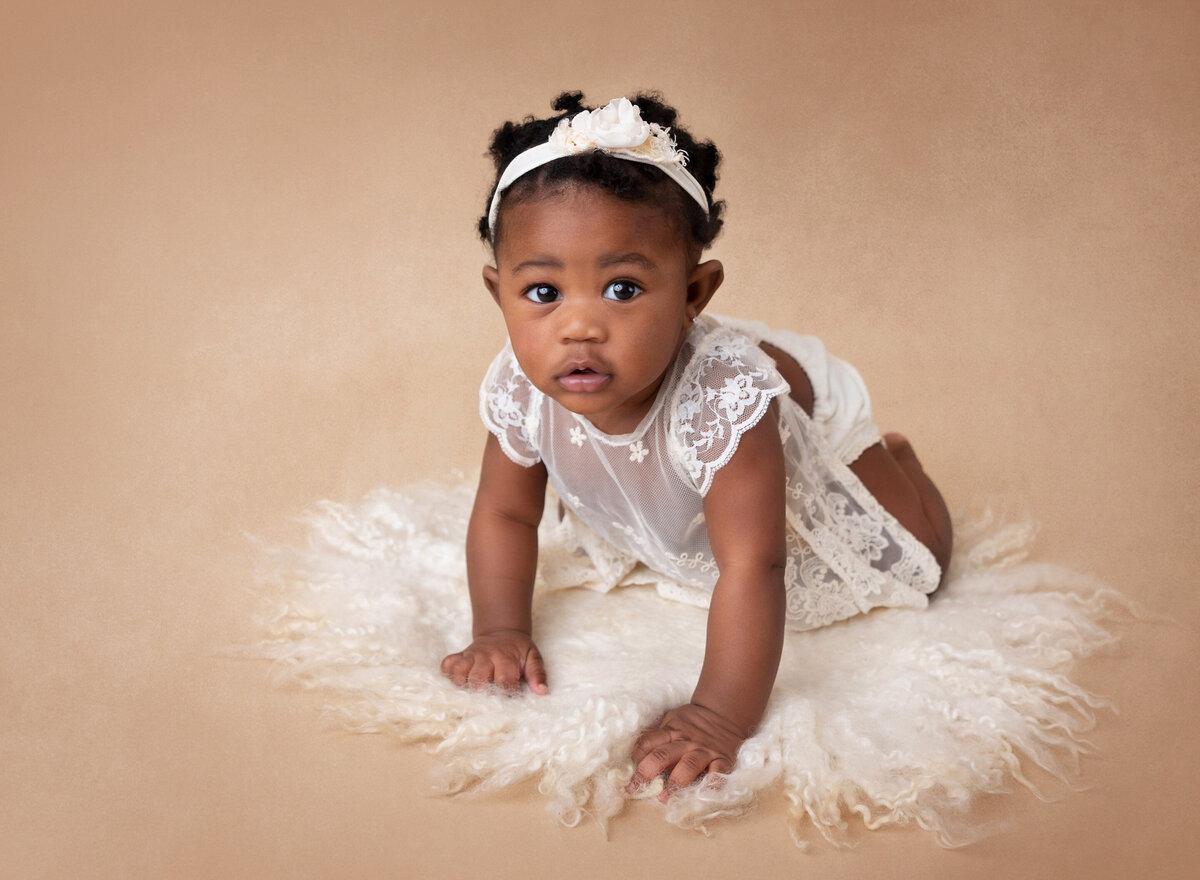Baby girl is crawling for a 6-month milestone photoshoot. She is wearing a white lace romper and looking at the camera.