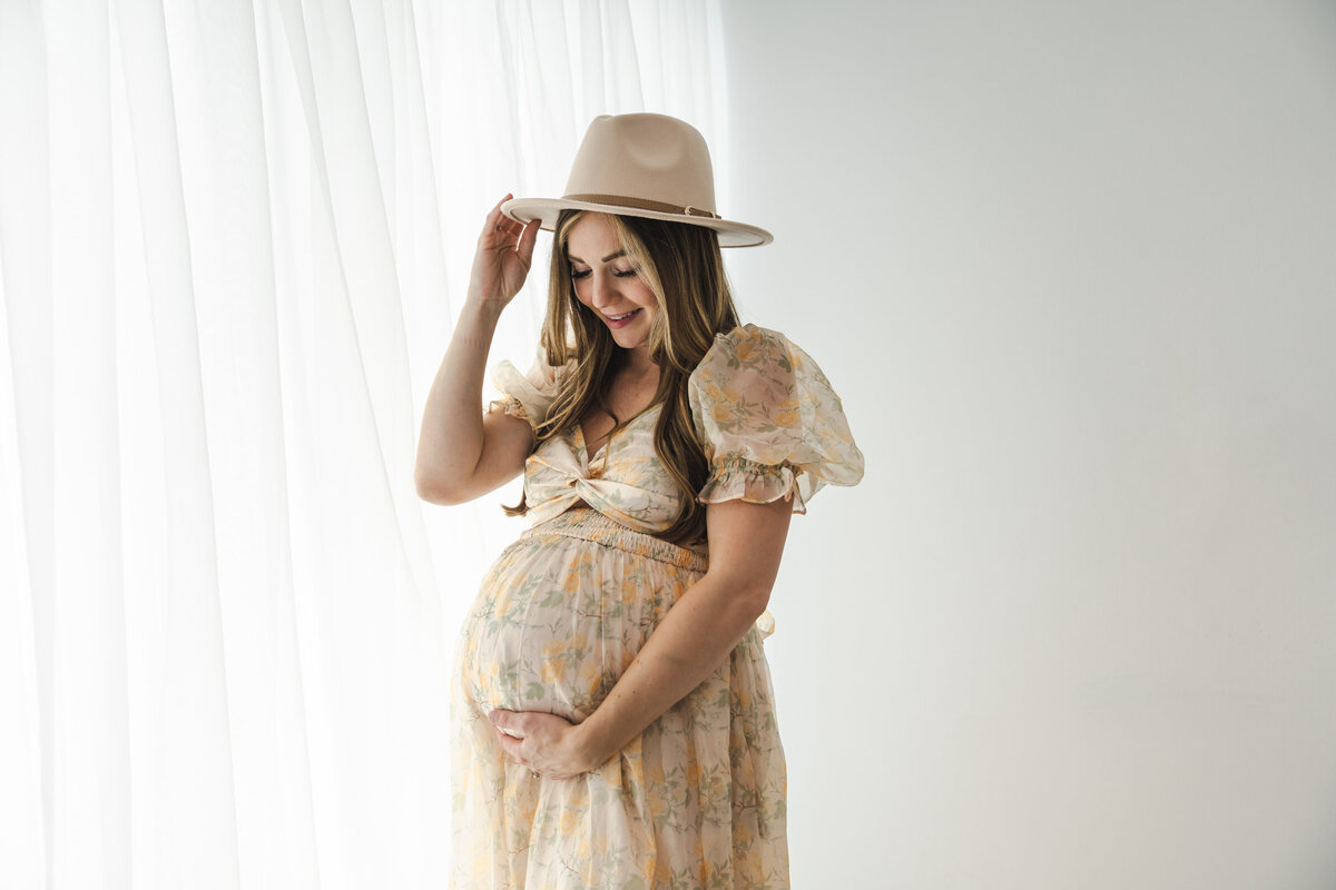 A smiling pregnant woman in a floral dress tenderly holds her belly while adjusting a stylish hat, standing against a bright background with sheer curtains. Taken by Fig and Olive Photography, a Minneapolis Maternity Photographer.
