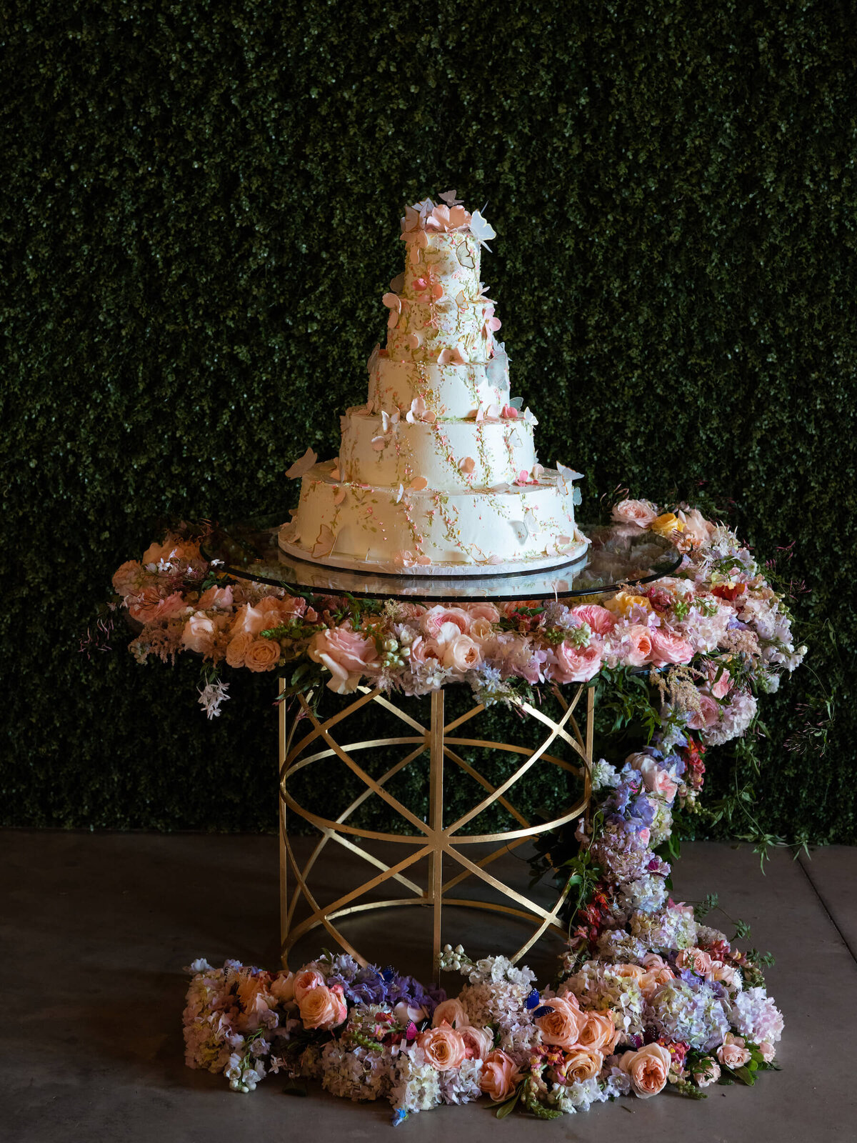 Pastel wedding cake covered in sugar flowers and edible butterflies sitting on floral cake table.