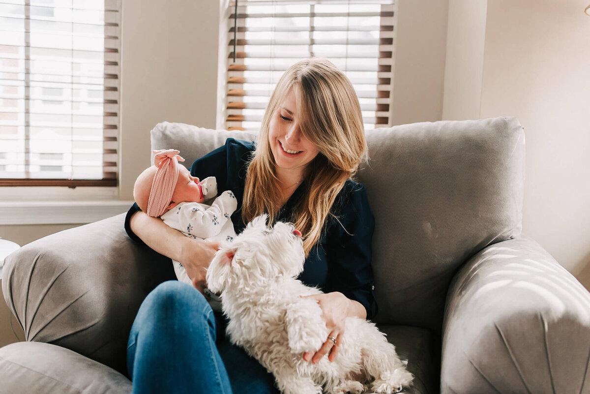 mom smiles and pets her dog while holding her baby girl on their grey recliner
