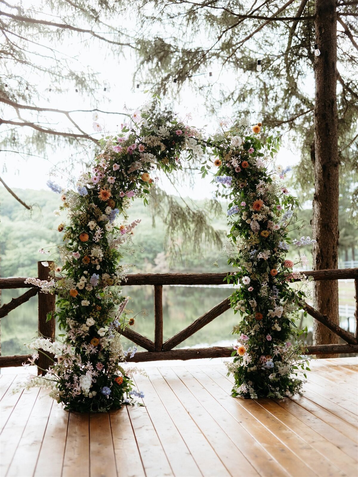 Stunning floral arbor by Canvas Weddings sits on wooden dock overlooking lake and trees at Cedar Lakes Estate wedding venue in Catskills