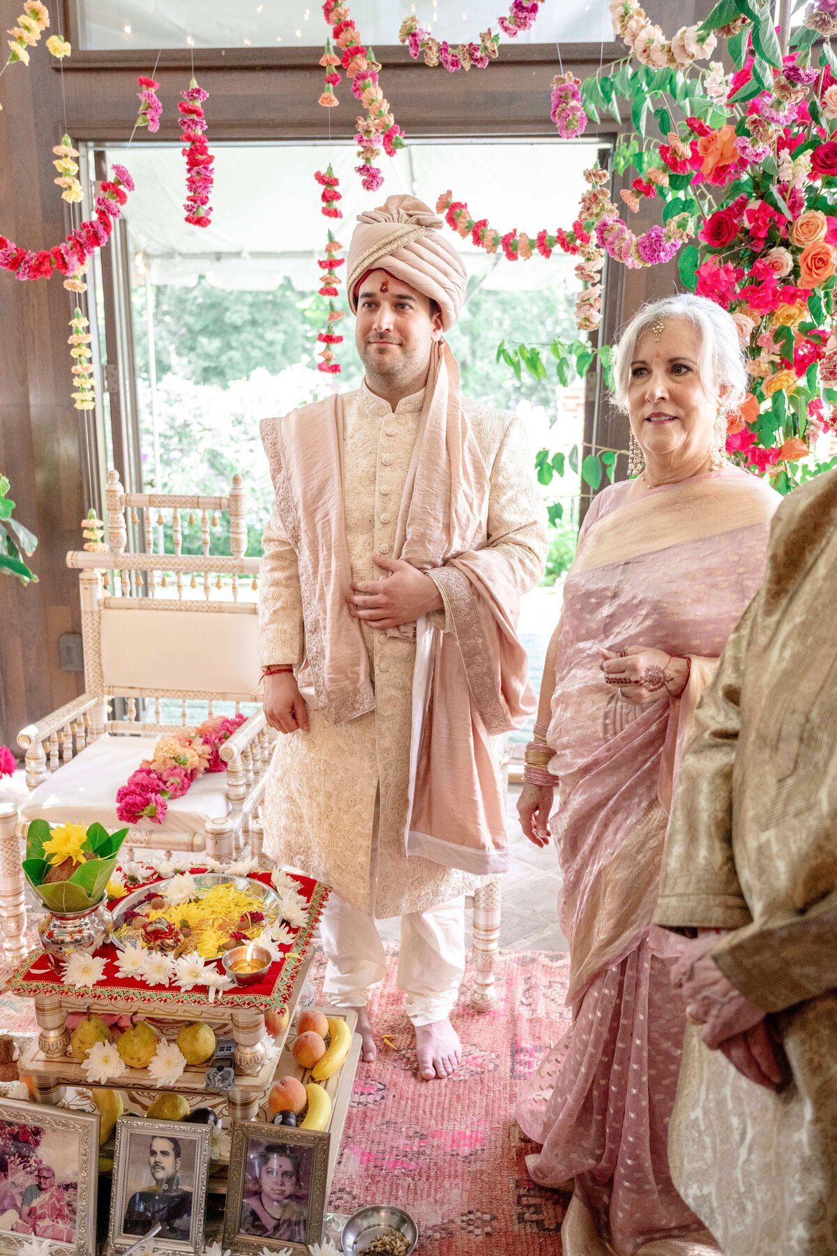 A groom in a traditional sherwani and turban stands beside an older woman in a pink saree. They are at a decorated ceremonial setting with flowers and a portrait.