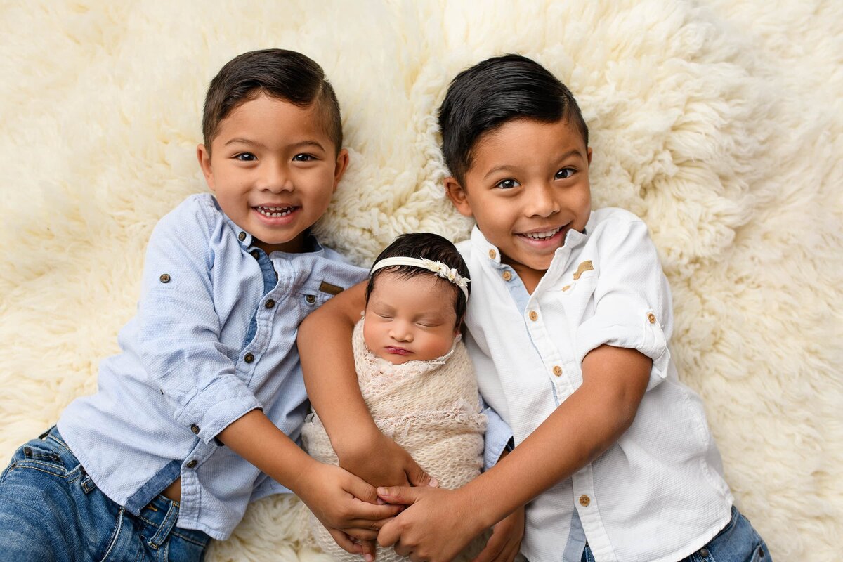 borthers snuggle their  newborn sister between them posed on a cream background