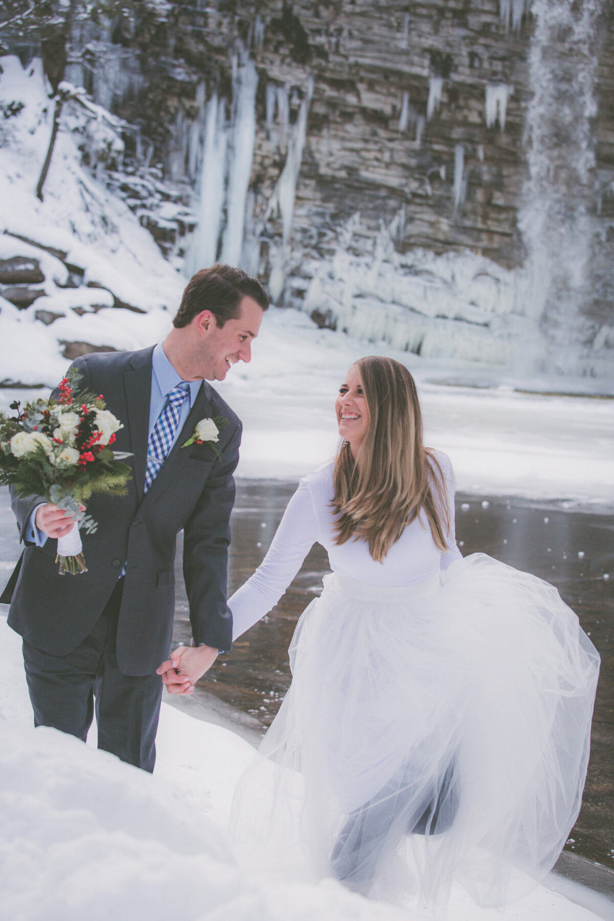Groom helps hold bouquet while couple walks through snow.