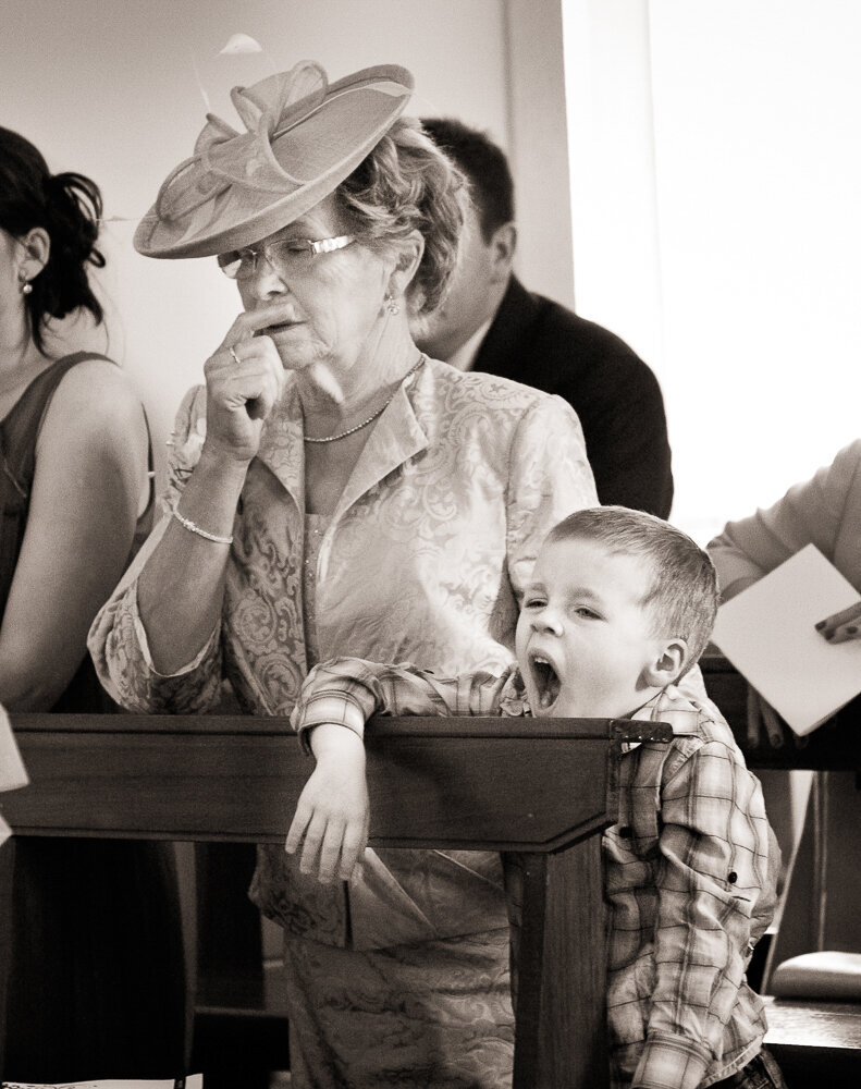 Mother of the bride wearing a fascinator hat sitting beside her grandson who is yawning in the church