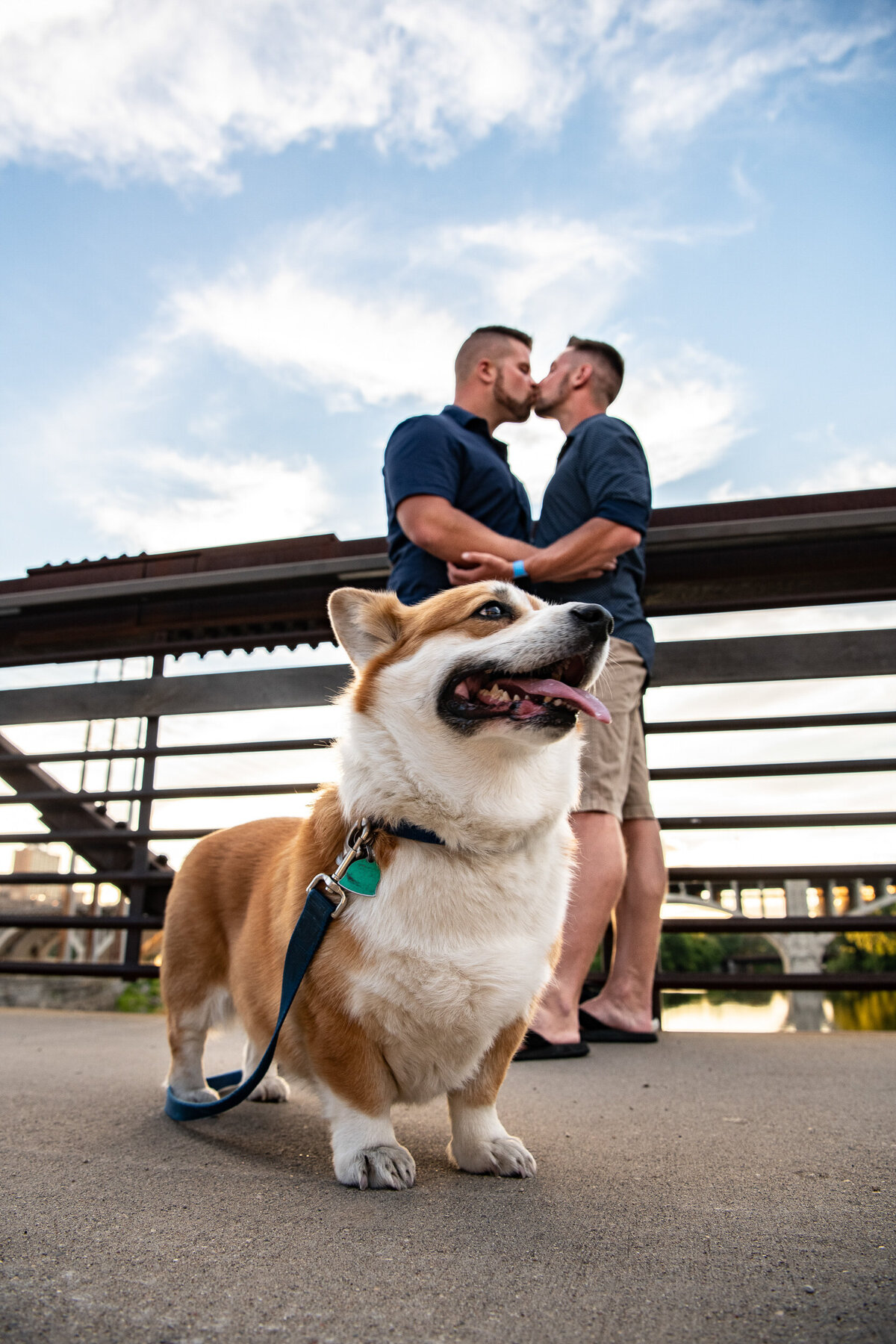 Two men kiss on a bridge behind their Corgi.