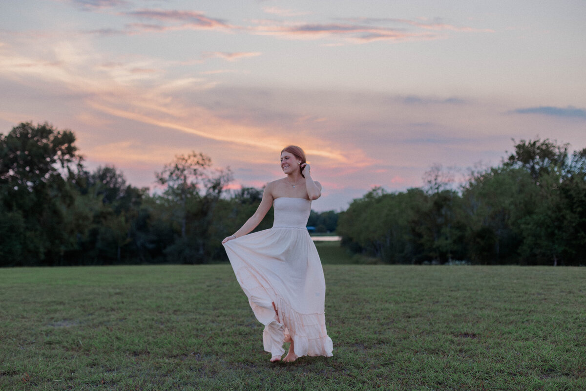 A senior girl runs in a green meadow while holding her maxi dress at sunset with a cotton candy sky