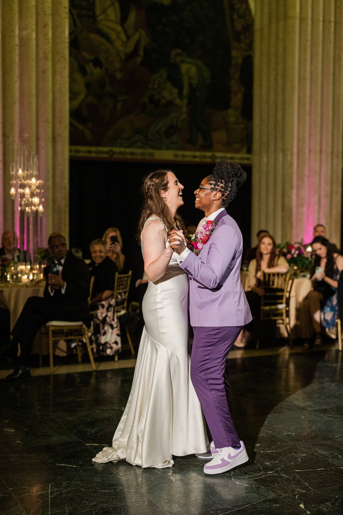 A candid photo of two brides happily sharing their first dance during their wedding reception at The Hall of State in Fair Park in Dallas, Texas. The bride on the left is wearing a long, white dress while the bride on the right wearing a purple suit with an large, intricate boutonniere and purple sneakers. Wedding guests can be seen watching intently from their tables in the background.