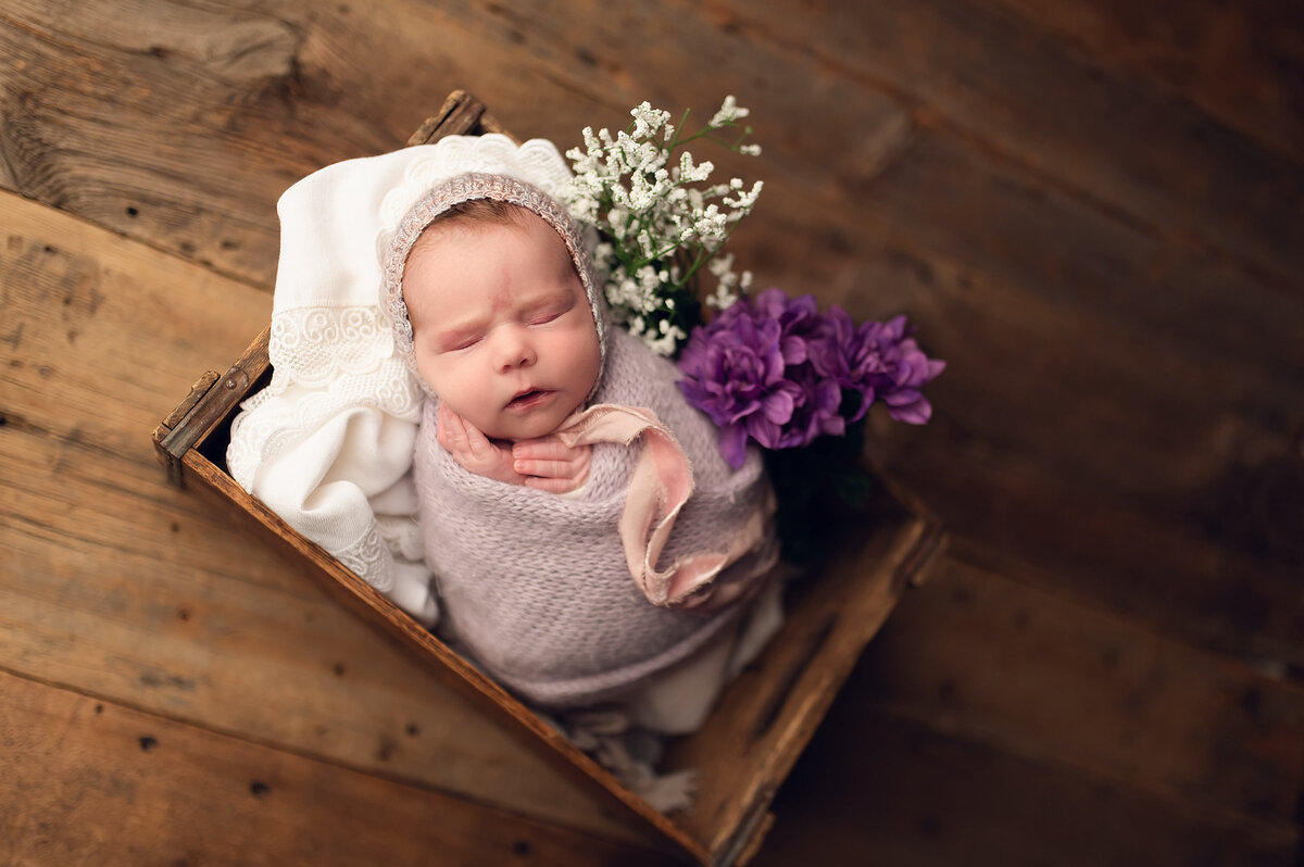 Portrait of a newborn baby asleep in a wooden crate swaddled in a lavender, woven blanket. Purple and white flowers  are placed along side.