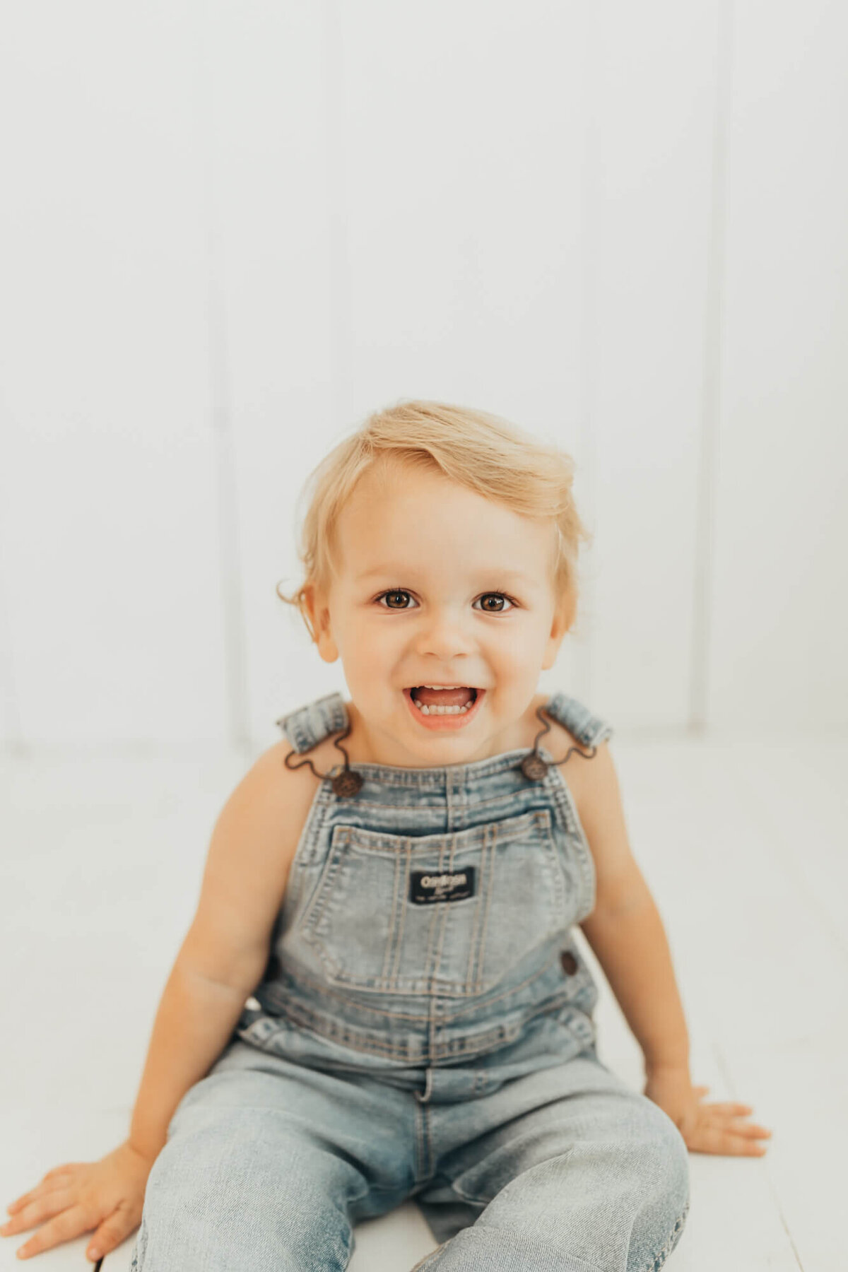 toddler boy smiles at the camera, wearing denim overalls.
