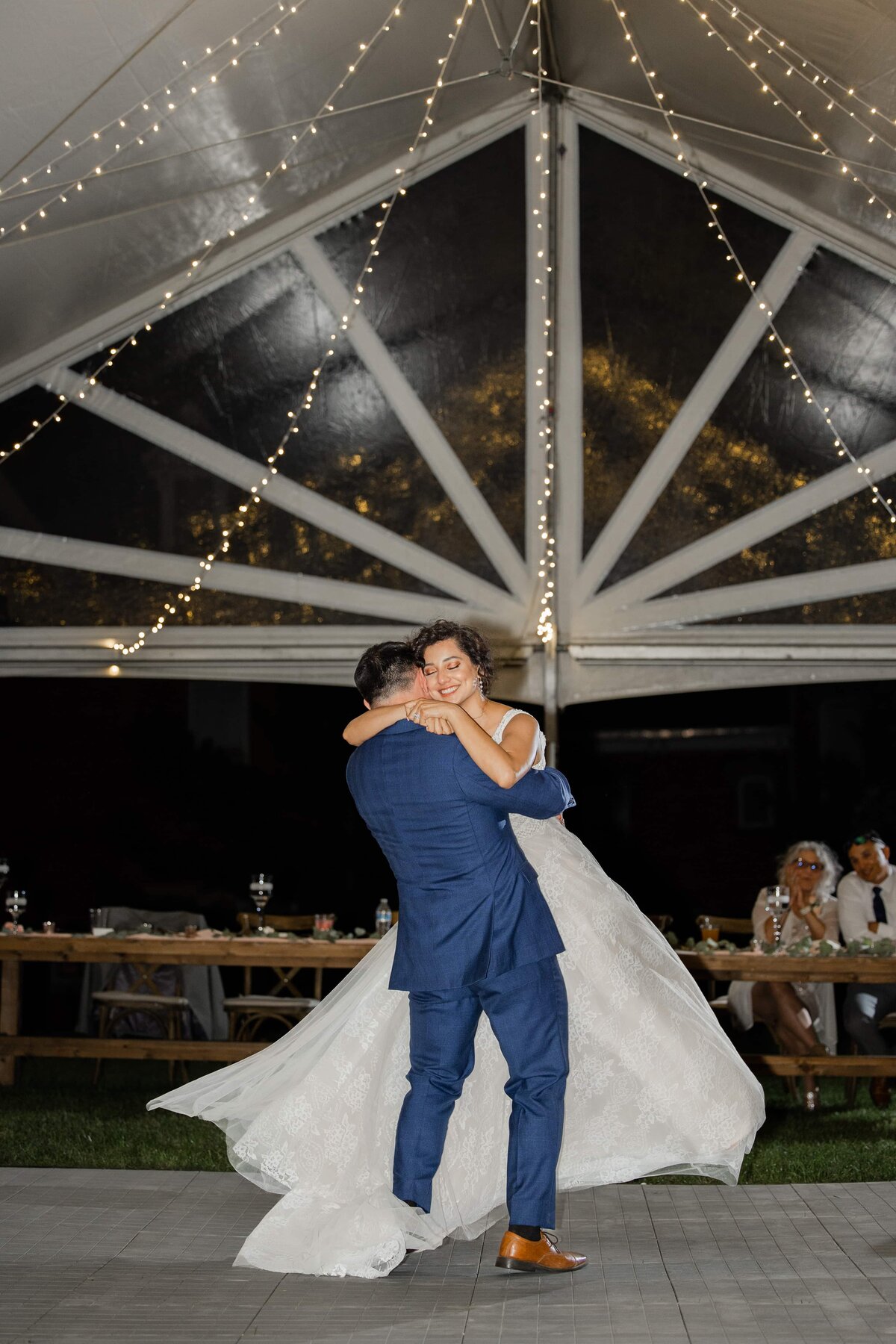 A bride and groom dance under a tent with string lights at a night-time wedding reception at a park farm winery, with guests seated around them.