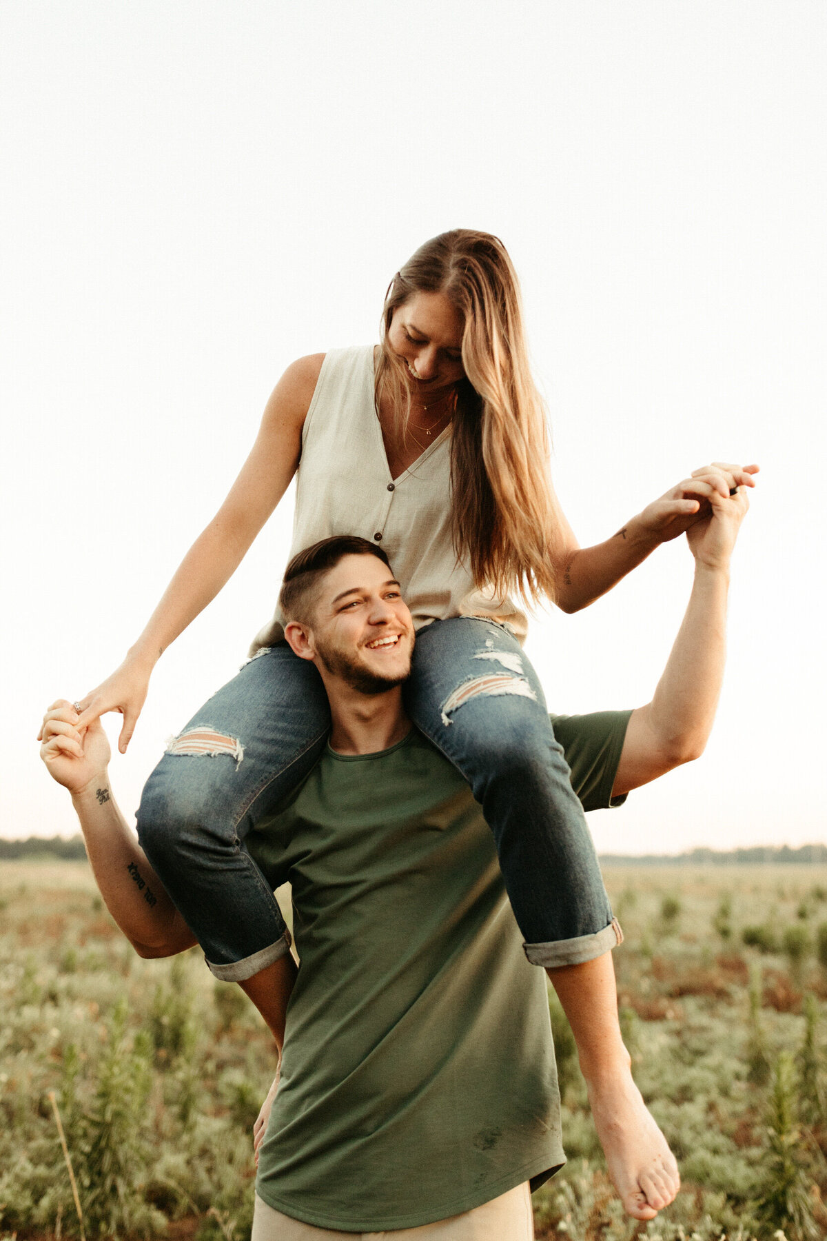 A girl is on top of a guy's shoulders as they hold hands and laugh together in a field.