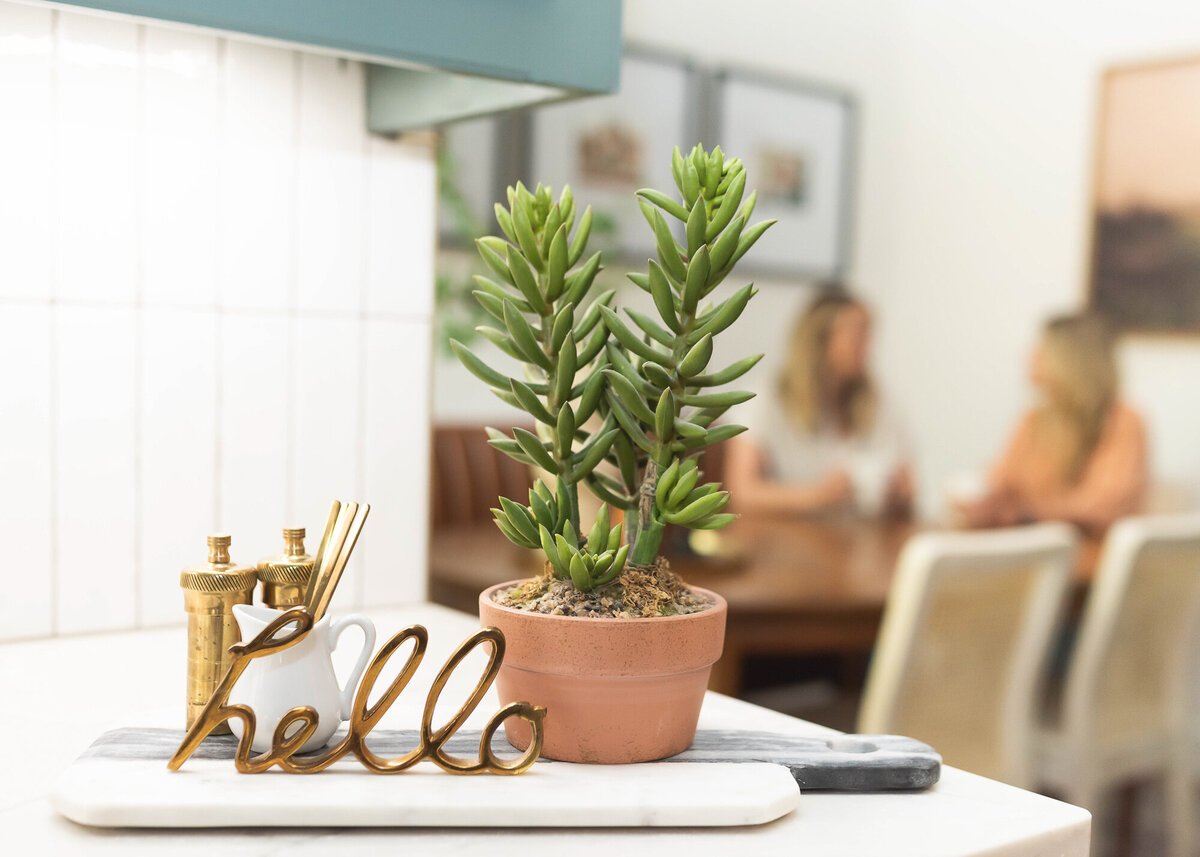 branding session content details shot of decor on counter that says hello, along with a small plant, and the owners of the company having coffee at the table behind, blurred out