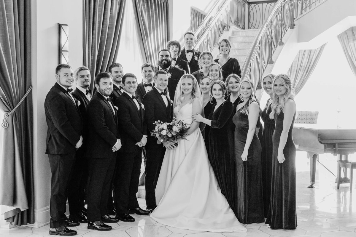 A black and white photo of a joyful Iowa wedding group, featuring a bride and groom with their bridesmaids and groomsmen, posed on and beside a grand staircase.