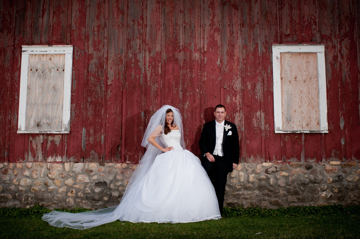A bride in a cinderella dress and groom pose for a portrait in front of a red barn