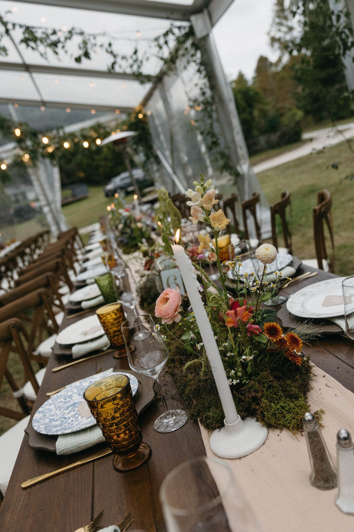 Close-up of reception table centerpiece featuring delicate garden-style wedding flowers including ranunculus, snapdragon, and wildflower-inspired stems arranged on a moss runner with taper candles and amber glassware. Designed by Fleurish Floral Studio for a whimsical outdoor wedding at The Nest in Northwest Arkansas.