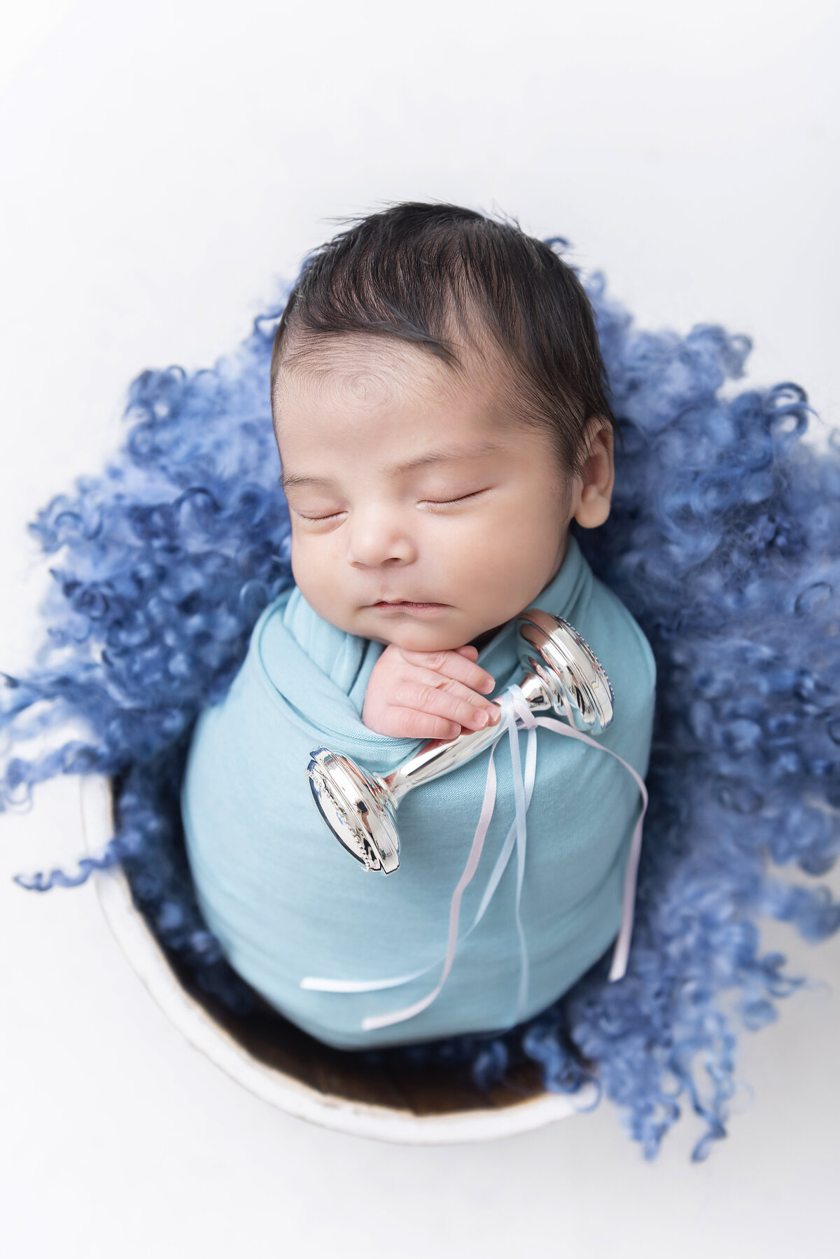 A newborn baby sleeps in a wooden bucket and blue blanket holding a tiny metal rattle posed by an Atlanta newborn photographer