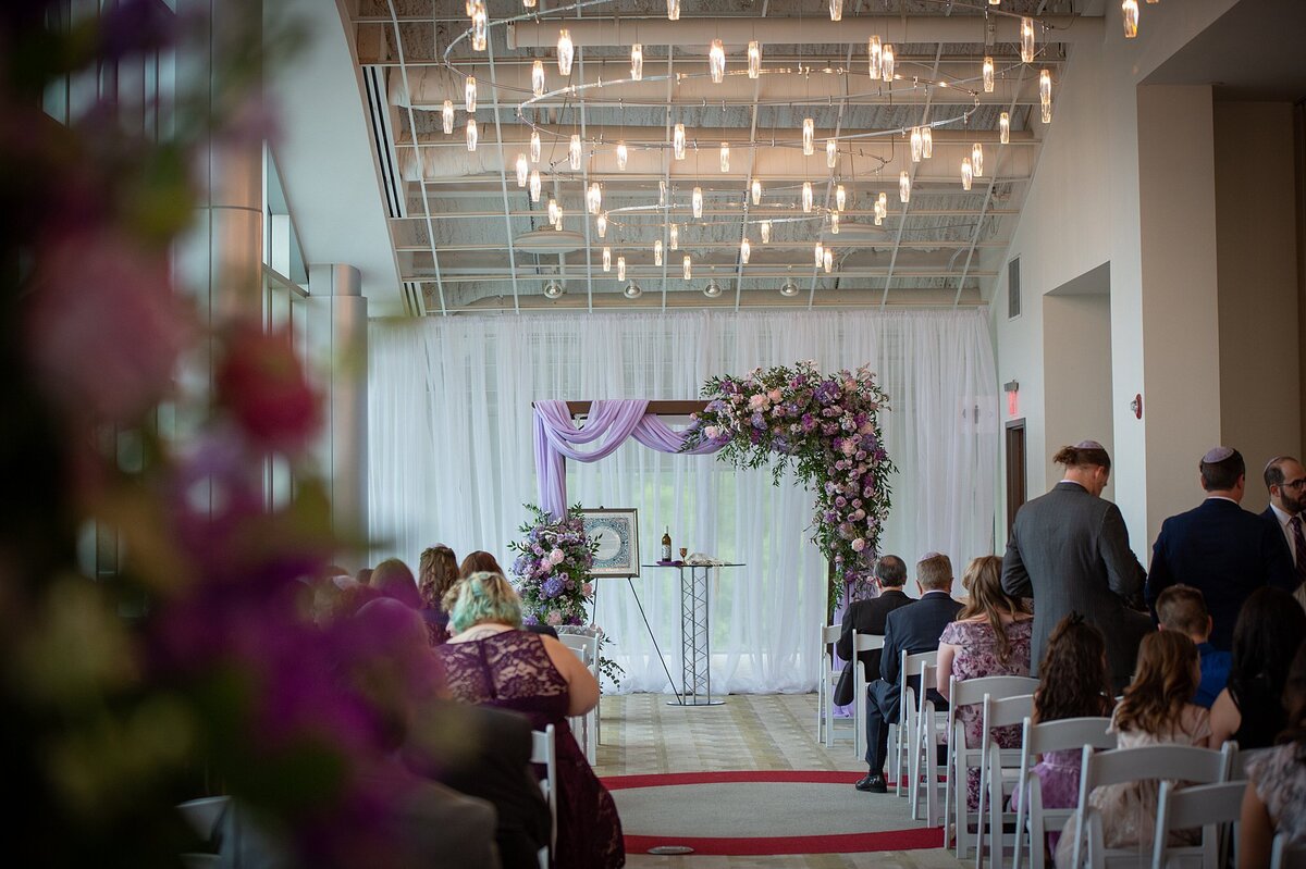 Wooden chuppah draped with with sheer purple fabric and decorated with an extra large spray of  purple roses, pink roses, white hydrangea and light purple roses. A glass cocktail table sits under the chuppah with a kiddish cup and bottle of wine next to the blue and white ketubah as guests are seated in white garden chairs at Noah Liff Opera Center.