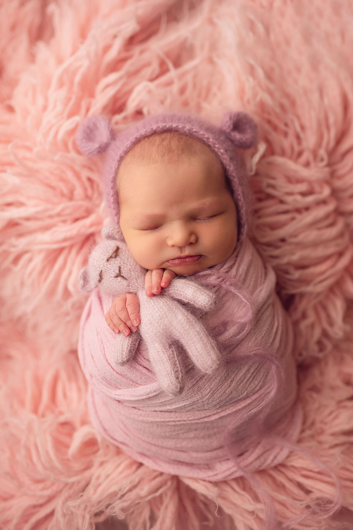 Tiny baby girl poses, sleeping, in our Waukesha studio. She is swaddled in pink muslin laid on a pink , shag rug while clutching a tiny pink teddy bear in her hands.