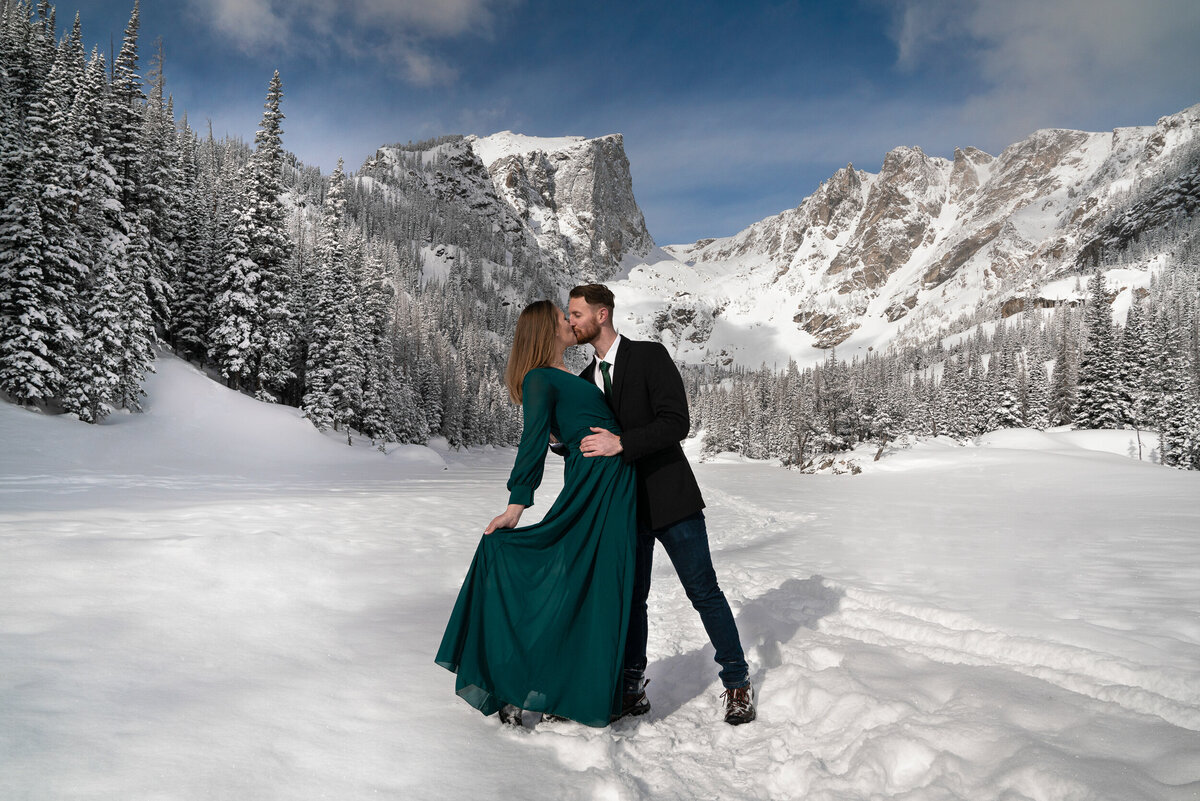 Man in suit and woman in green dress kiss in front of the Rocky Mountains.