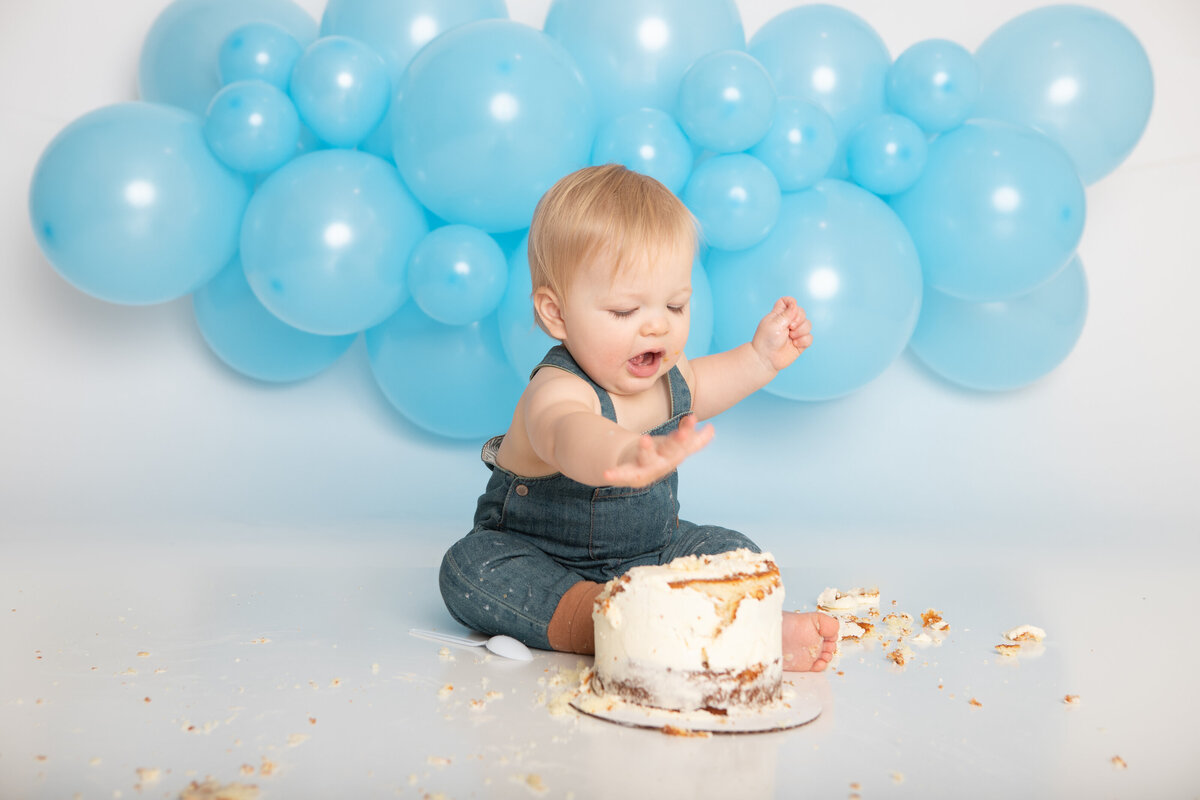 Baby eating a messy white cake with a blue balloon garland