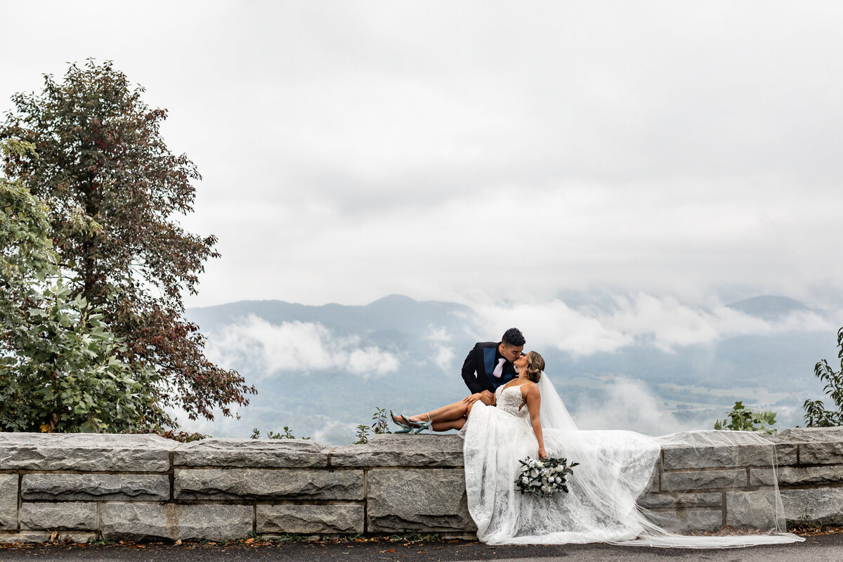 Bride on a wall with her legs out and dress on on the ground while the groom leans over and kisses her. The fog and Smoky Mountains are in the background. Capturing Love Amidst the Smoky Peaks