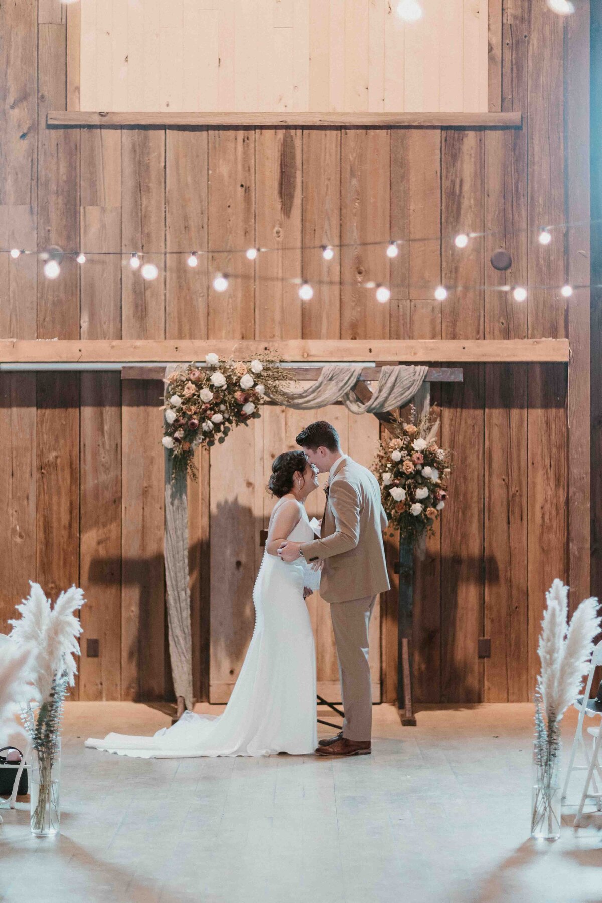 Bride and groom in a tan suit stand under their wooden arbor and market lights
