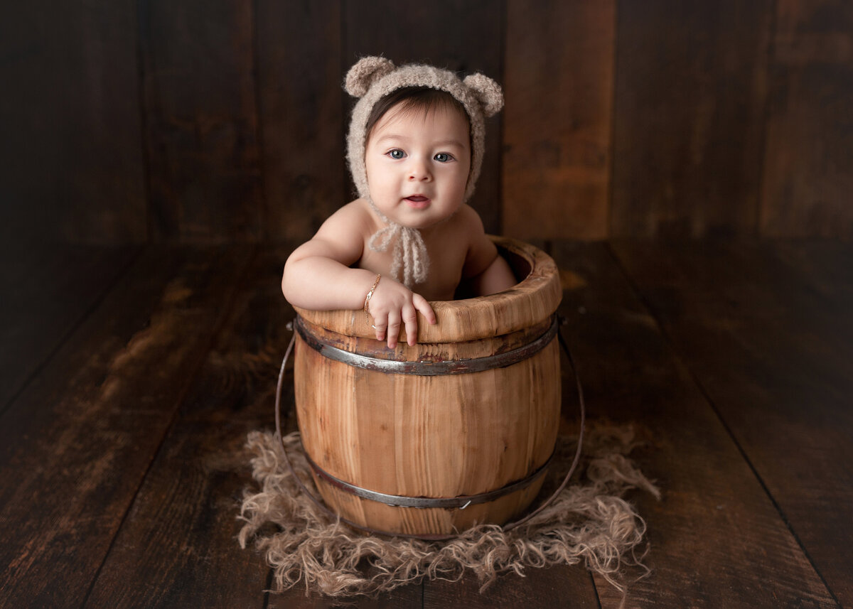 12-month baby photoshoot  at West Palm Beach and Lake Worth photography studio. Baby is sitting in a wooden bucket with his hand on the edge. Baby is wearing a fuzzy bear bonnet. Bucket is placed on a wood infinity backdrop.