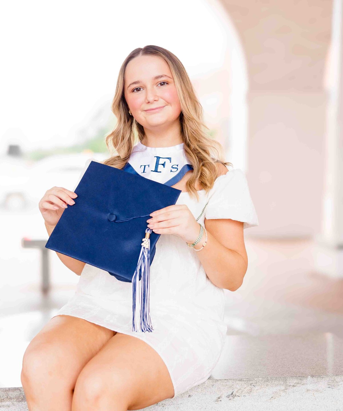 A young woman sits outdoors, holding a blue graduation cap with a white and blue tassel. Dressed in a white dress and sporting a "TFS" stole, she is the focus against a softly blurred background. Captured by a senior photographer in Fayetteville, NC, her joy radiates through the lens.