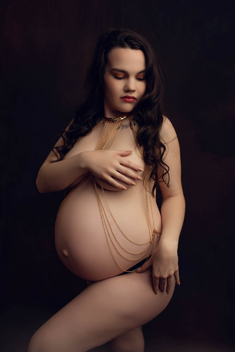 A mostly nude pregnant woman wearing a bodysuit covering her breasts with her hand
