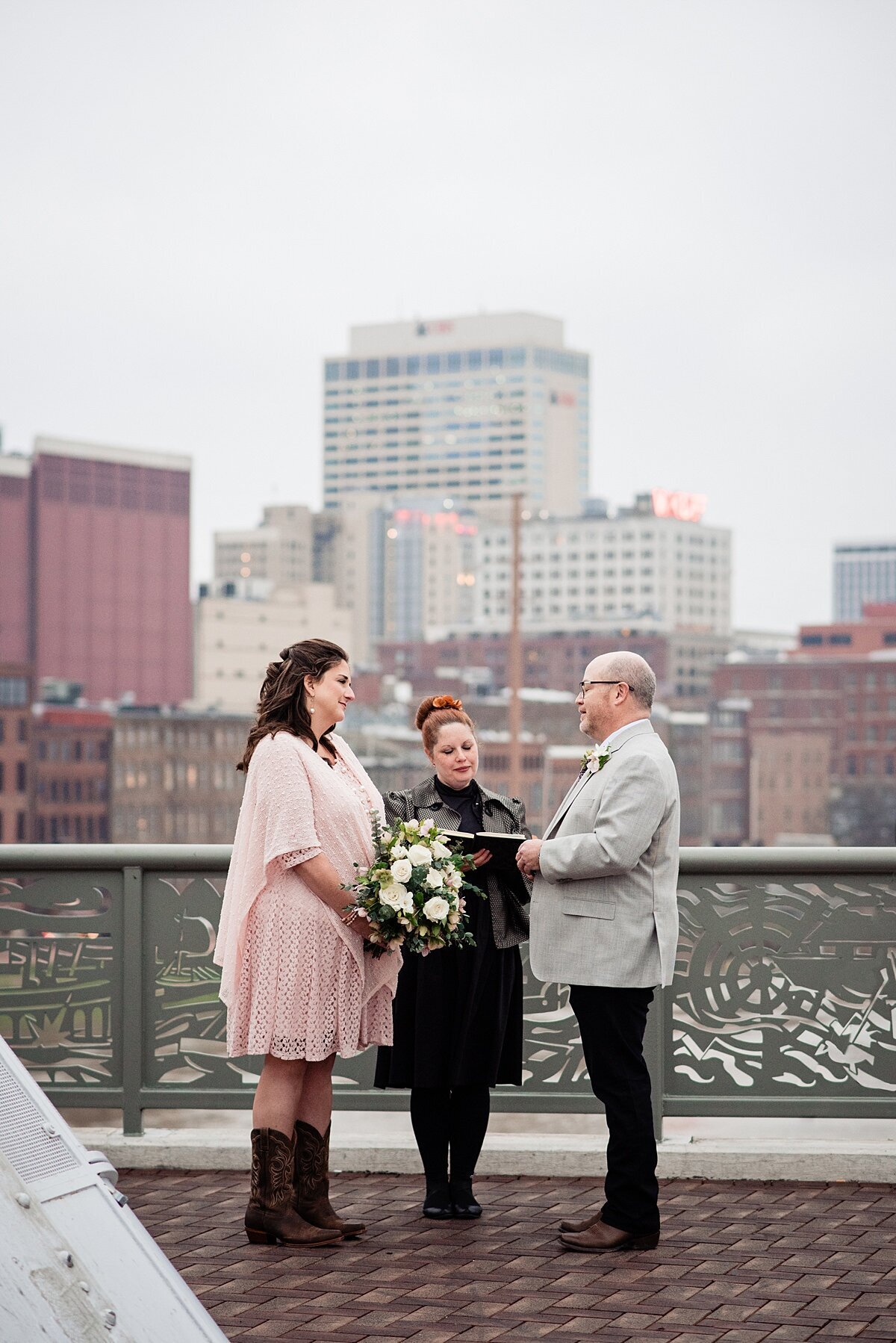 The bride and groom stand with their officiant on the Pedestrian Bridge in Nashville with the Nashville Skyline behind them. The bride is wearing a flowing knee length gauze  boho dress with elbow length sleeves and brown cowboy boots. The groom is wearing a light gray jacket and black pants. The bride is holding a large white bouquet  as she and the groom smile at each other. The wedding officiant is wearing a black dress with black tights. Her red hair is up in a structured bun. She is reading from a leather bound book as they take their vows.