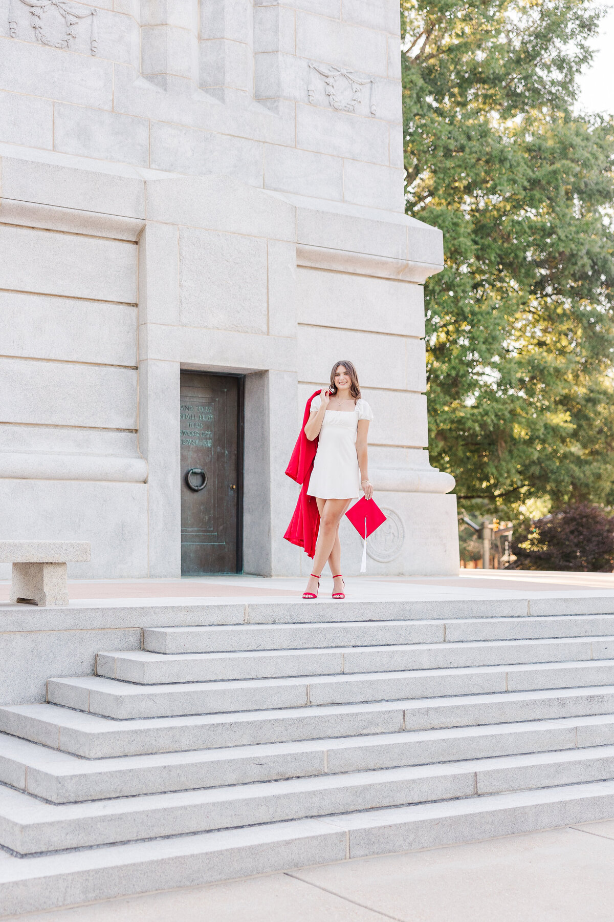 A woman in a white dress stands gracefully on stone steps in front of a large white building, expertly captured by a senior photographer in Fayetteville, NC. She holds a red jacket and wears matching red high heels, with elegant trees framing the background.