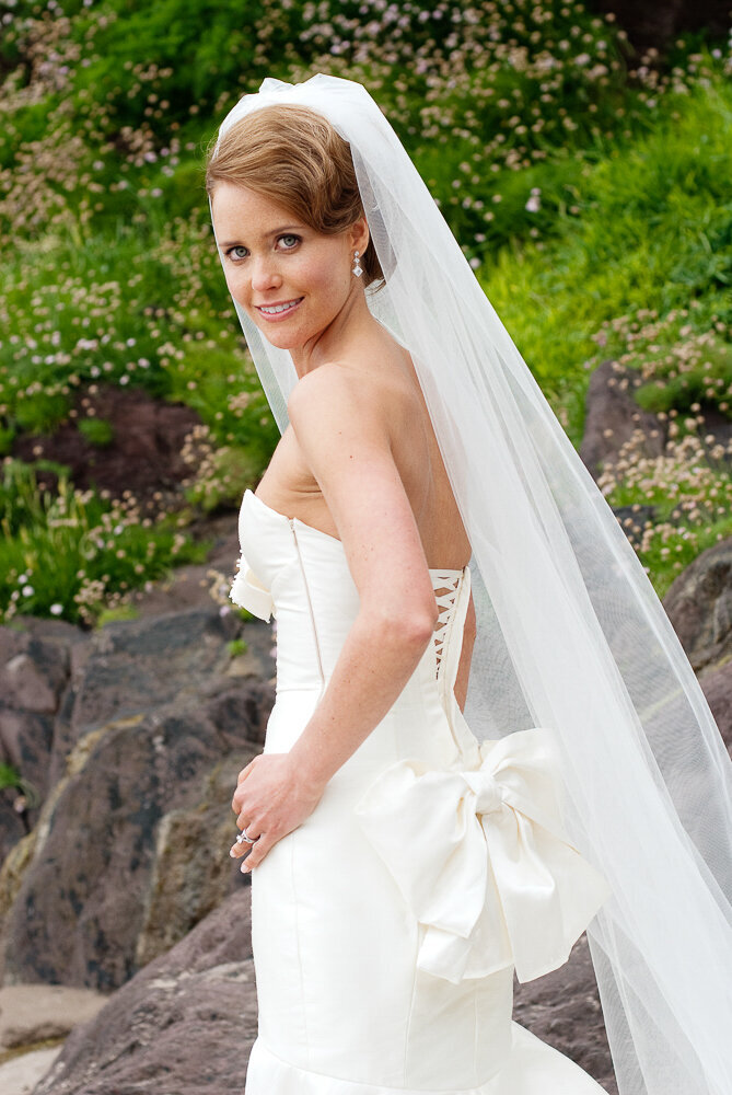 bride wearing a mermaid style wedding dress with corset bodice and large bow with long veil looking over her shoulder while standing on a beach