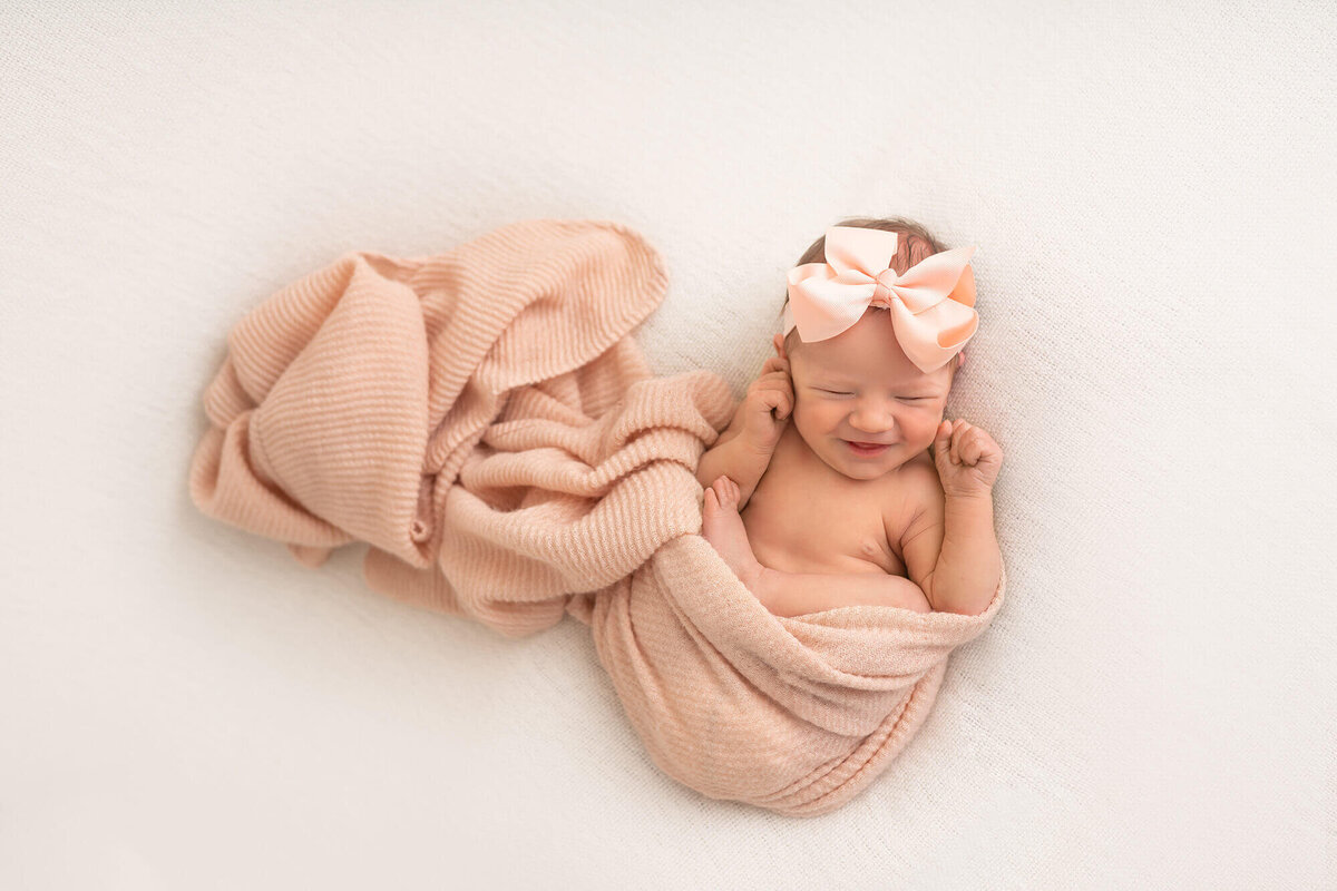 A newborn baby girl wrapped in a pink blanket and wearing a giant pink bow smiles while sleeping on a white blanket