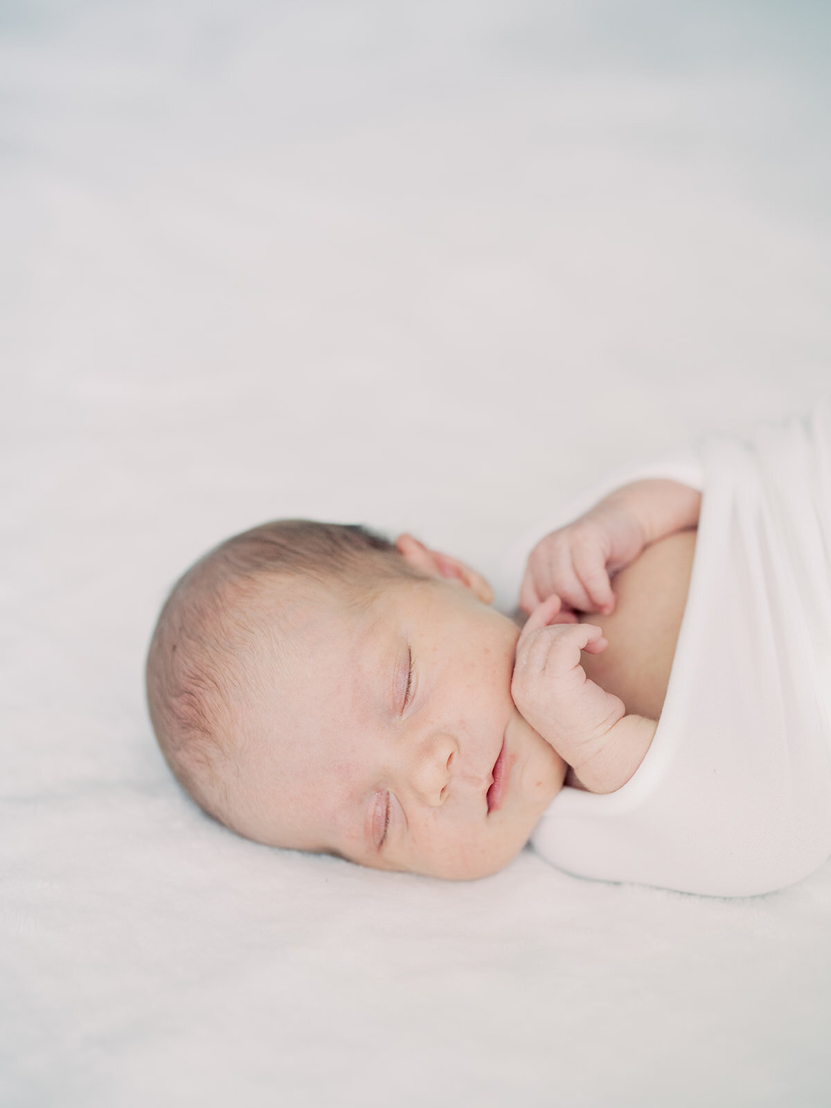 Newborn baby swaddled in white sleeps peacefully on a bed.