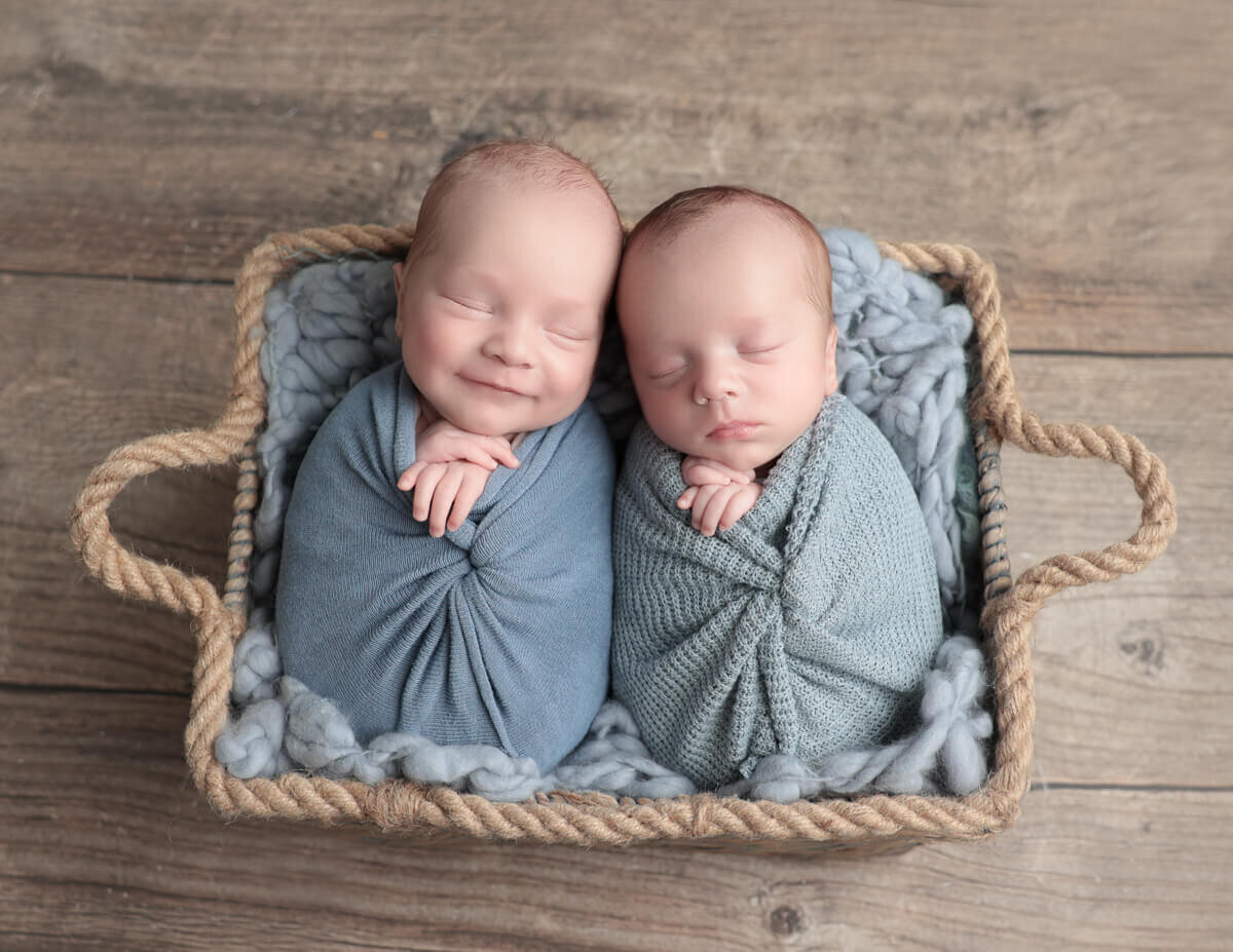 A charming photo of twin newborn boys wrapped in soft blue blankets, peacefully nestled in a woven basket. Captured in a cozy photography studio in Rochester, NY, this image showcases the beauty of twin newborn photography with delicate details and gentle lighting.