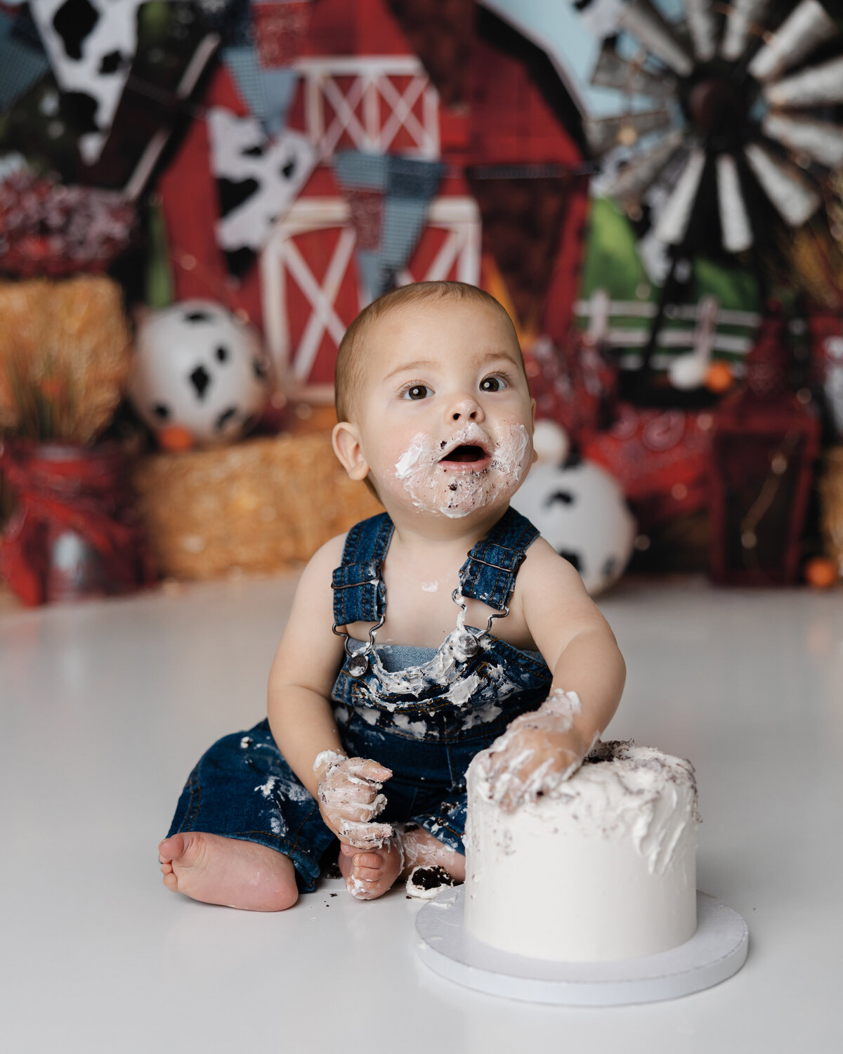 First birthday portrait of a baby boy in denim overalls, sitting in front of a red barn backdrop with cake in hand. Photograph by Yucaipa cake smash photographer Kaitlyn Dawn Photography.