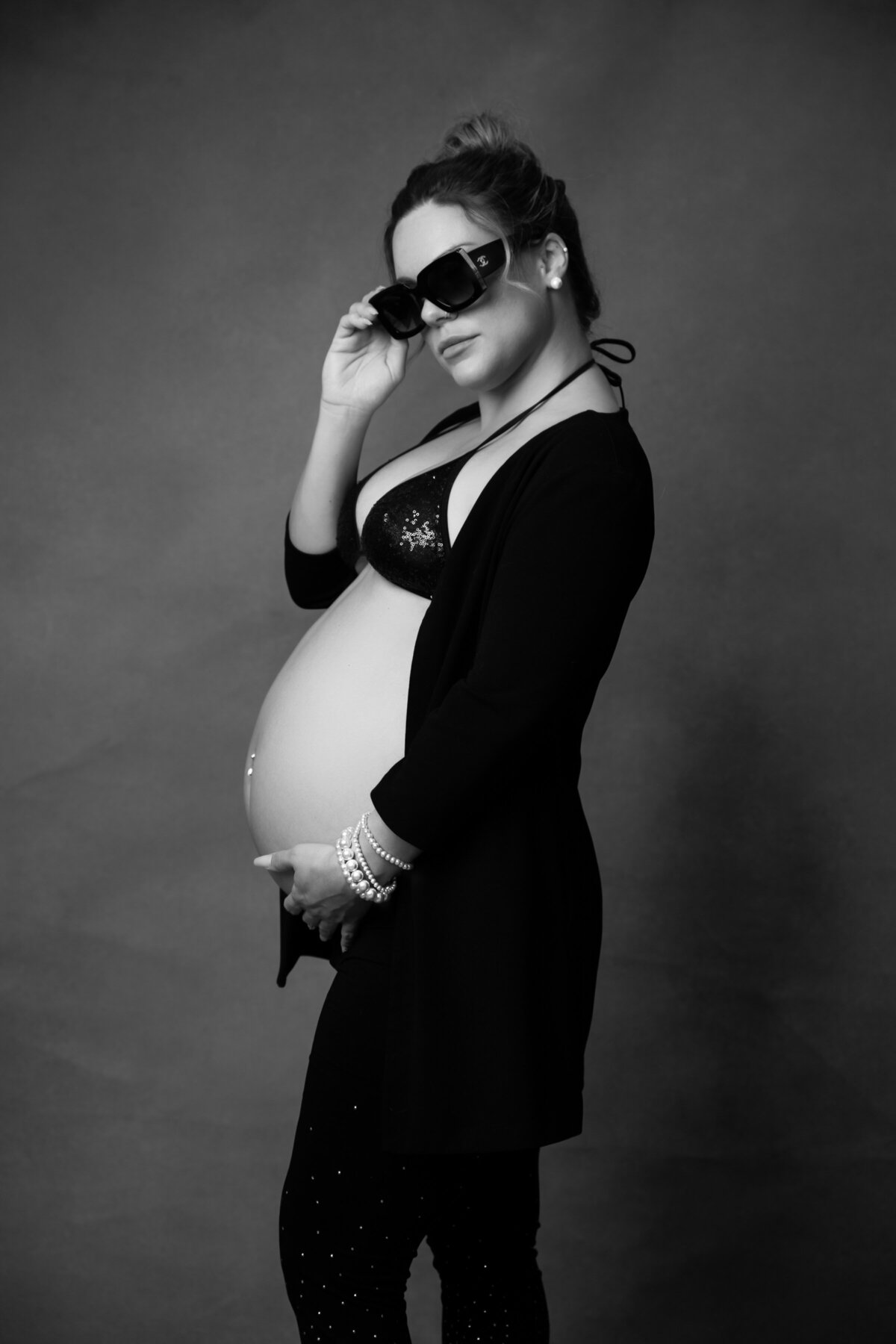 Black-and-white portrait of a stylish pregnant woman posing confidently against a dark backdrop. She is wearing oversized sunglasses, a sequined bikini top, a long open cardigan, and pearl bracelets, exuding a bold, glamorous vibe. Her hand rests on her belly, highlighting her silhouette and the strength and elegance of motherhood with a touch of Hollywood flair.