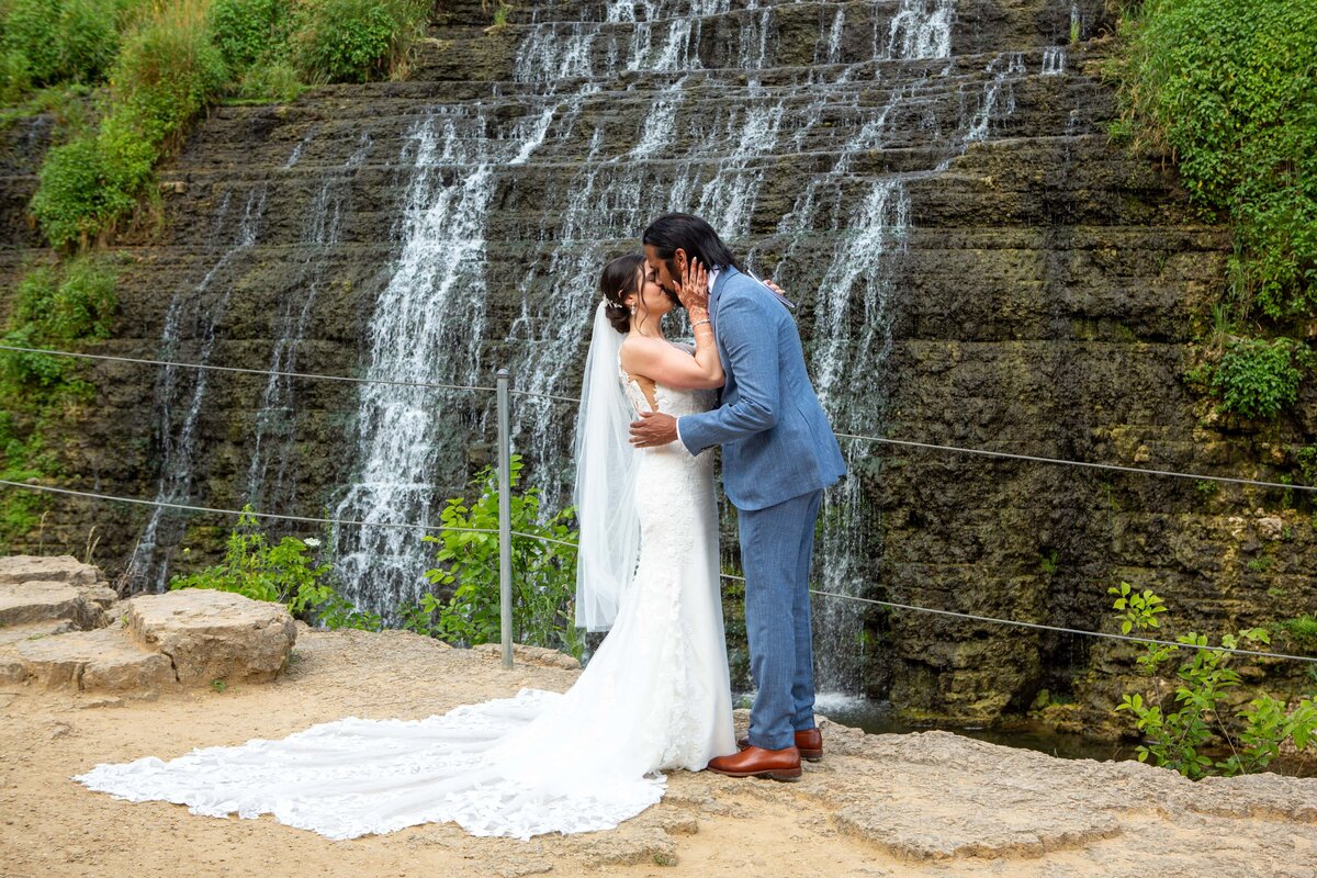 A bride in a white gown and a groom in a blue suit kiss in front of a small waterfall with lush greenery, coordinated by a top wedding planner from Des Moines.